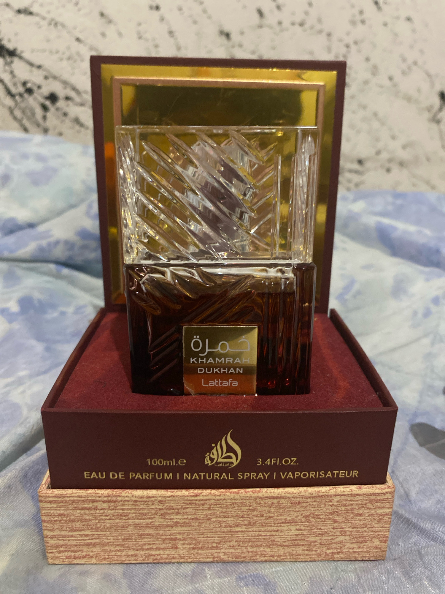 The image shows a close-up of a perfume bottle and its packaging, set against a blurred background of fabric. The perfume bottle has a dark amber liquid and a clear, faceted glass cap. A gold label on the bottle reads "KHAMRAH DUKHAN" and "Lattafa". The bottle is housed in a dark red presentation box. The exterior of the box has text that includes "100ml.e", "3.4FI.OZ.", "EAU DE PARFUM | NATURAL SPRAY | VAPORISATEUR", and a logo. The packaging rests on a patterned surface that appears to be part of a bedspread or blanket. The image lacks any discernible details that would place it in the city of Barcellona Pozzo di Gotto, Italy.