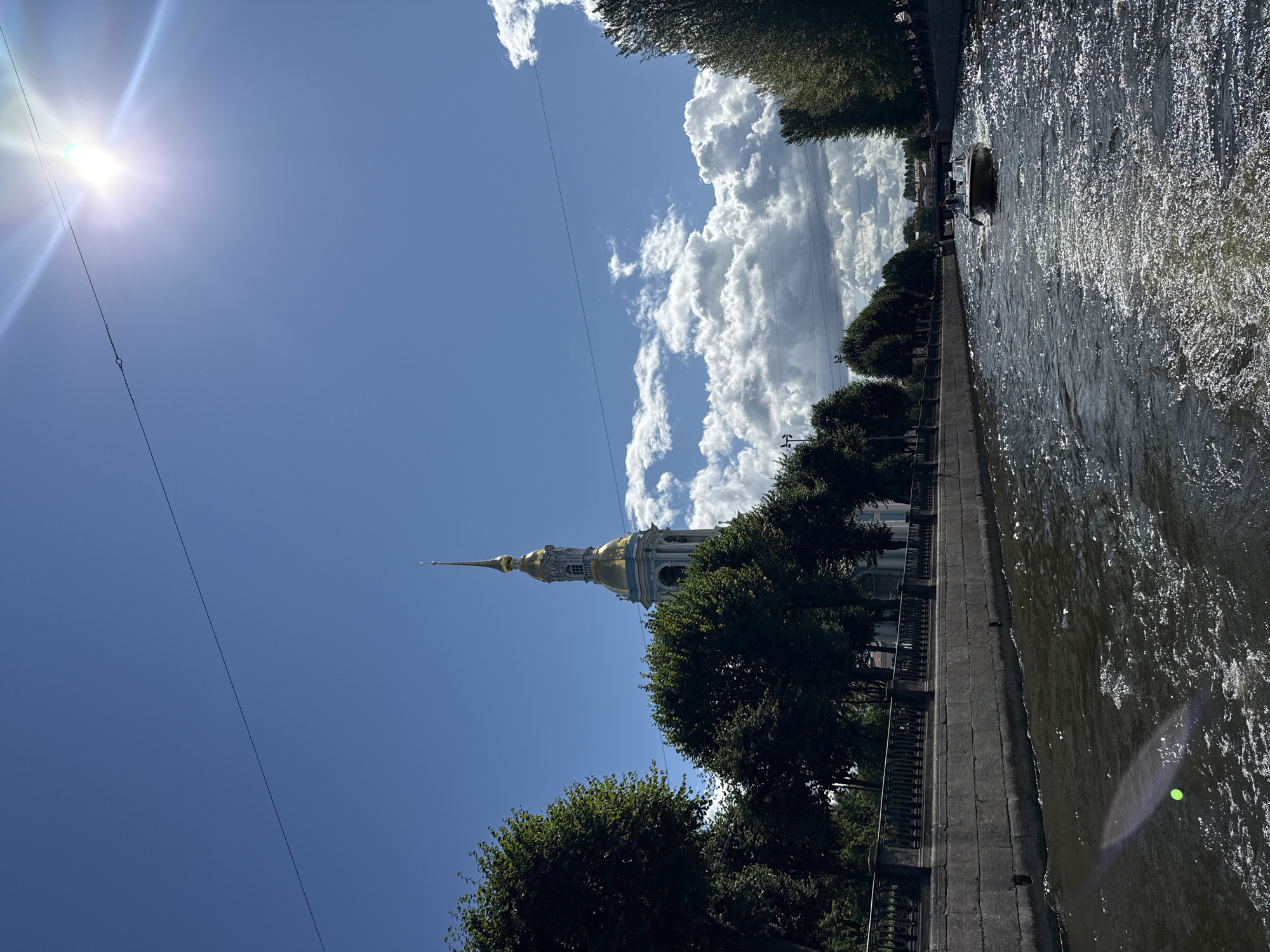 This is a photograph taken from a boat on a canal in Saint Petersburg, Russia. The canal is lined with stone embankments and trees. On the left side of the canal, a church spire with a golden dome rises above the trees. In the distance, a bridge crosses the canal. A small motorboat is visible in the middle of the canal, moving away from the viewer. The sky is blue with scattered white clouds, and the sun is shining brightly, creating reflections on the water. The overall impression is a sunny day in a European city with historical architecture.