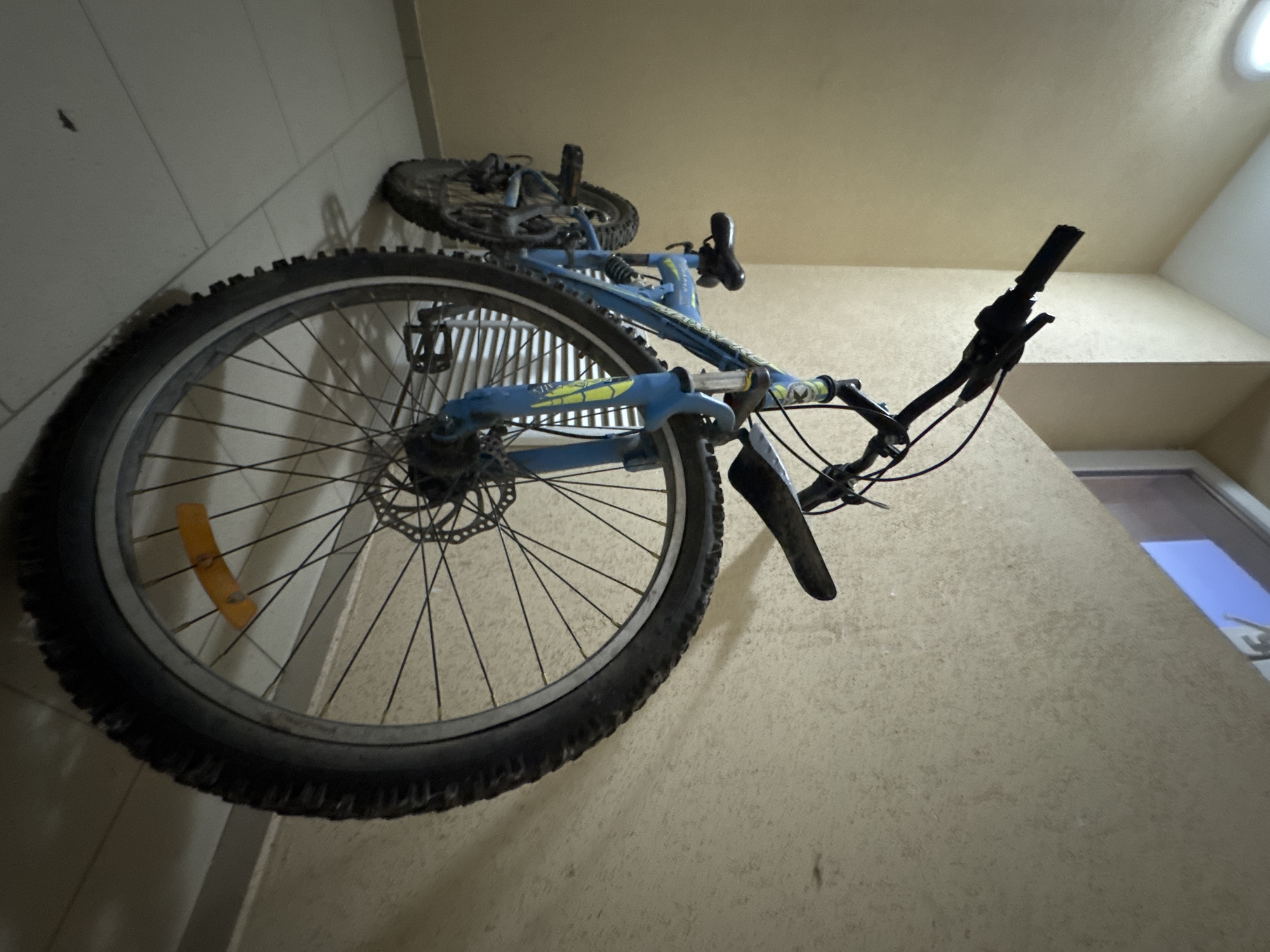 The image shows a blue and black bicycle propped up against a wall. The bicycle is oriented vertically, with its front wheel facing the viewer and its rear wheel at the top of the frame. The bicycle appears to be a mountain bike, with knobby tires and suspension. The frame is primarily blue, with yellow and black accents.
The background is a textured, light-colored wall. The setting appears to be indoors, possibly a garage or storage area, given the way the bike is propped and the lighting. There are no people visible in the image.
Notable details include a yellow reflector on the spokes of the front wheel. The time of day is indeterminate due to the indoor lighting. No weather conditions can be inferred. No visible text is present on the bicycle or in the background. The overall impression is one of a functional bicycle stored against a wall. The location context of Mikhaylovsk, Russia, provides geographical information but does not add specific visual cues to the image itself.