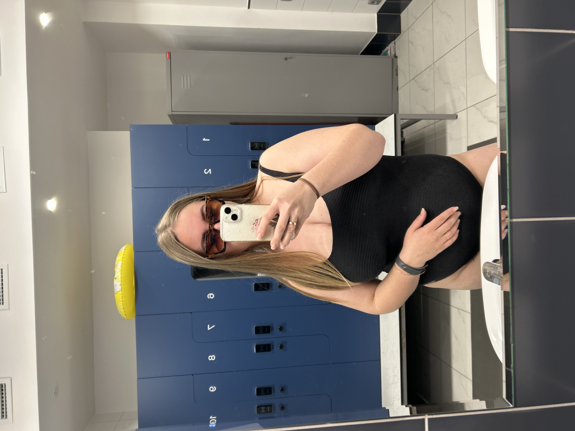 A medium shot shows a woman taking a mirror selfie in what appears to be a locker room. The woman has blonde hair and is wearing sunglasses. She's holding a smartphone in her right hand to take the picture and is wearing a black one-piece swimsuit. Her left hand rests on her stomach.
Behind her, there are rows of blue lockers with electronic locks. A yellow inflatable ring is visible near the top of the lockers. The locker room has white walls and a tiled floor. A gray metal locker is visible at the top right. The bathroom fixtures and black tiled walls indicate the photo was taken in the city of Chaikovsky, Russia.