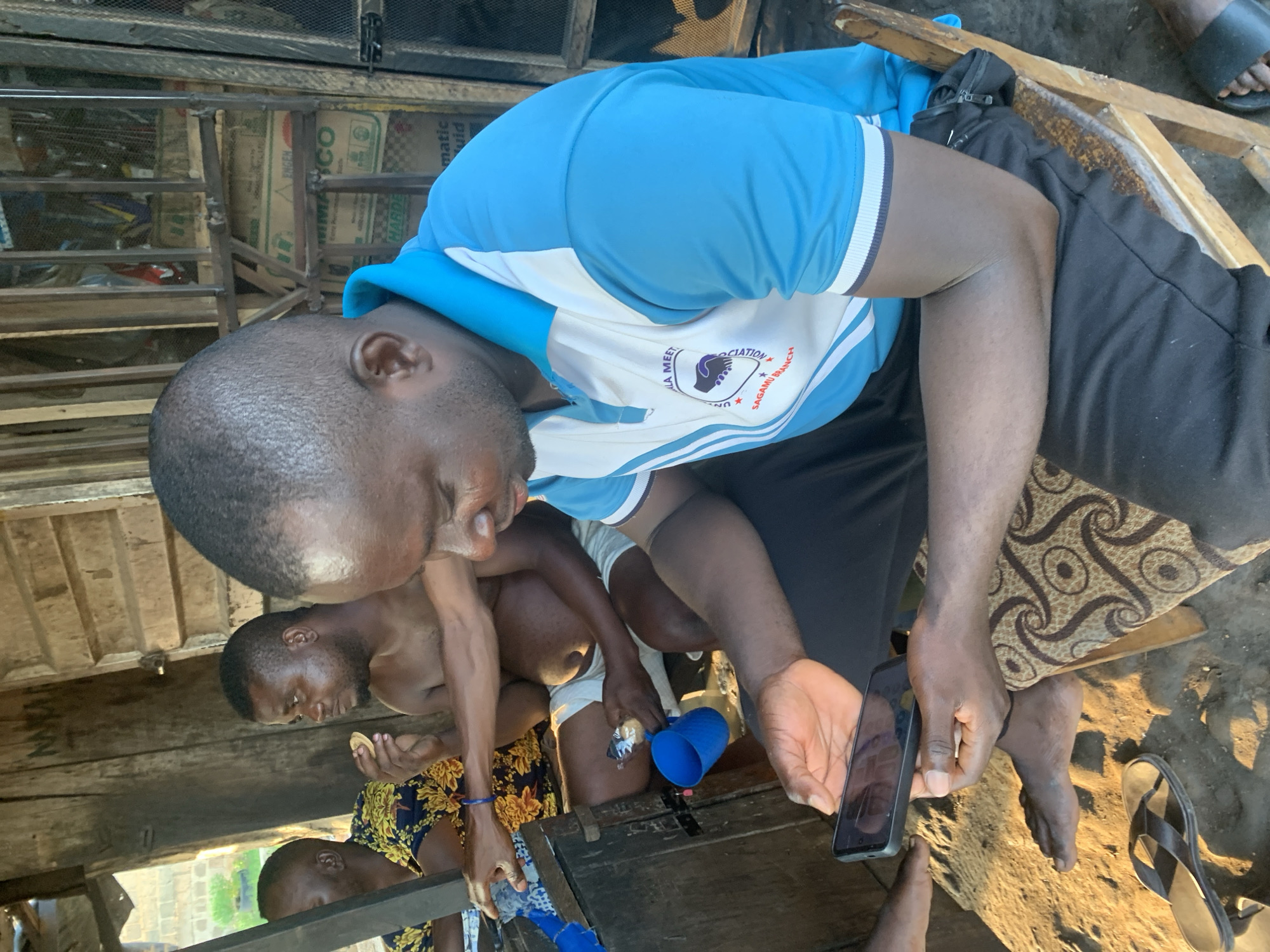 In Shagamu, Nigeria, four men are gathered under a rustic, open-sided structure with wooden beams and corrugated metal roofing. The man in the foreground, wearing a blue and white collared shirt with the inscription "SAGAMU BRANCH", is seated on a wooden chair and holding a smartphone, looking at its screen. To his left, another man, shirtless, is seated and holding a small, circular food item. A third man, partially visible on the far left, wears a colorful, patterned shirt. The fourth man is mostly out of frame. The ground is dirt and sand, with sandals visible near the front. The environment appears to be an outdoor gathering space, possibly a local shop or communal area, with various items and structures suggesting a functional but unrefined setting.