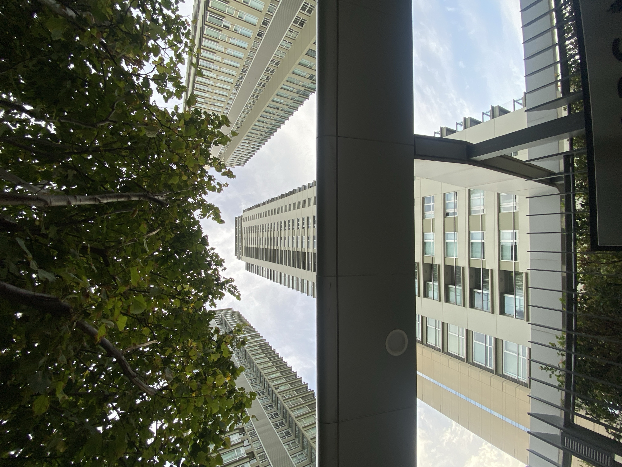 The image is an upward-angled view of buildings in an urban environment. The foreground contains portions of a building structure, including a grey pillar and what appears to be architectural details, with leafy trees partially obscuring the view on the left and right sides. In the background, tall buildings with rows of windows rise into the sky, which is partly covered with clouds. The precise location in Buenos Aires, Argentina, is not apparent from the image alone.