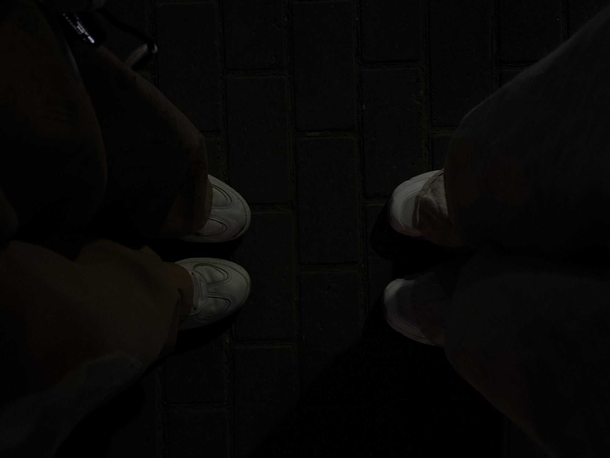 The image shows two pairs of legs and white sneakers standing on a dark, brick-paved surface. The location is potentially in the city of Perm, Russia, based on the image location (if any) which is not provided. The scene is dimly lit, making details about clothing and facial features (if visible) difficult to ascertain. The image is composed from a high-angle perspective, focusing on the lower portion of two individuals. The surrounding environment appears to be a walkway or public area with brick paving. There is no visible interaction or action occurring in the frame.