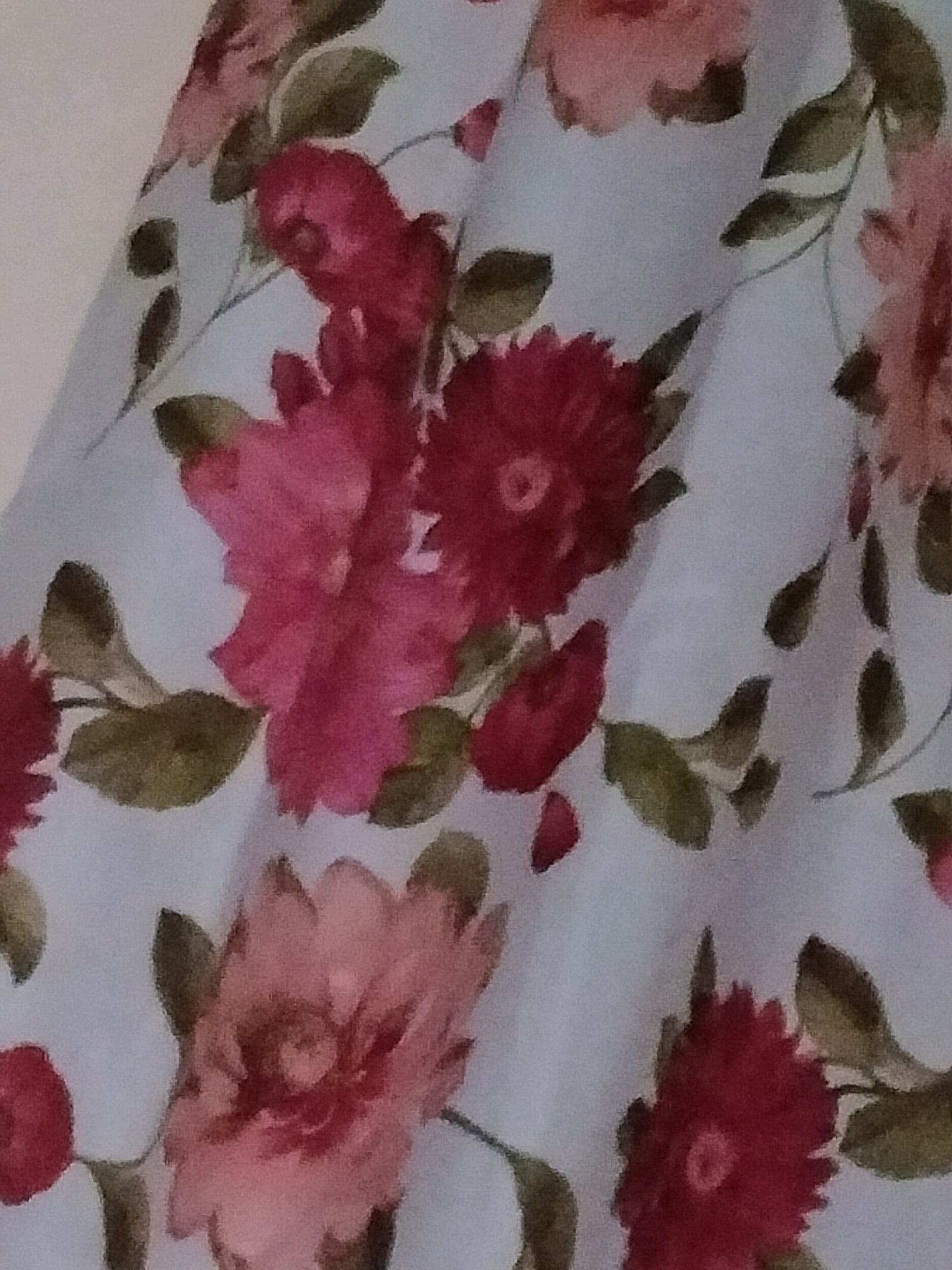 The image displays a close-up view of a light blue fabric with a floral pattern. The pattern consists of pink and deep red flowers with green leaves. The flowers appear to be roses or similar multi-petaled blooms. The fabric is draped, creating folds and shadows that add depth to the image. The lighting suggests an indoor setting. There are no people visible in the image. The location in Samad ash Shan, Oman, is not discernible from the visual content of the image itself.
