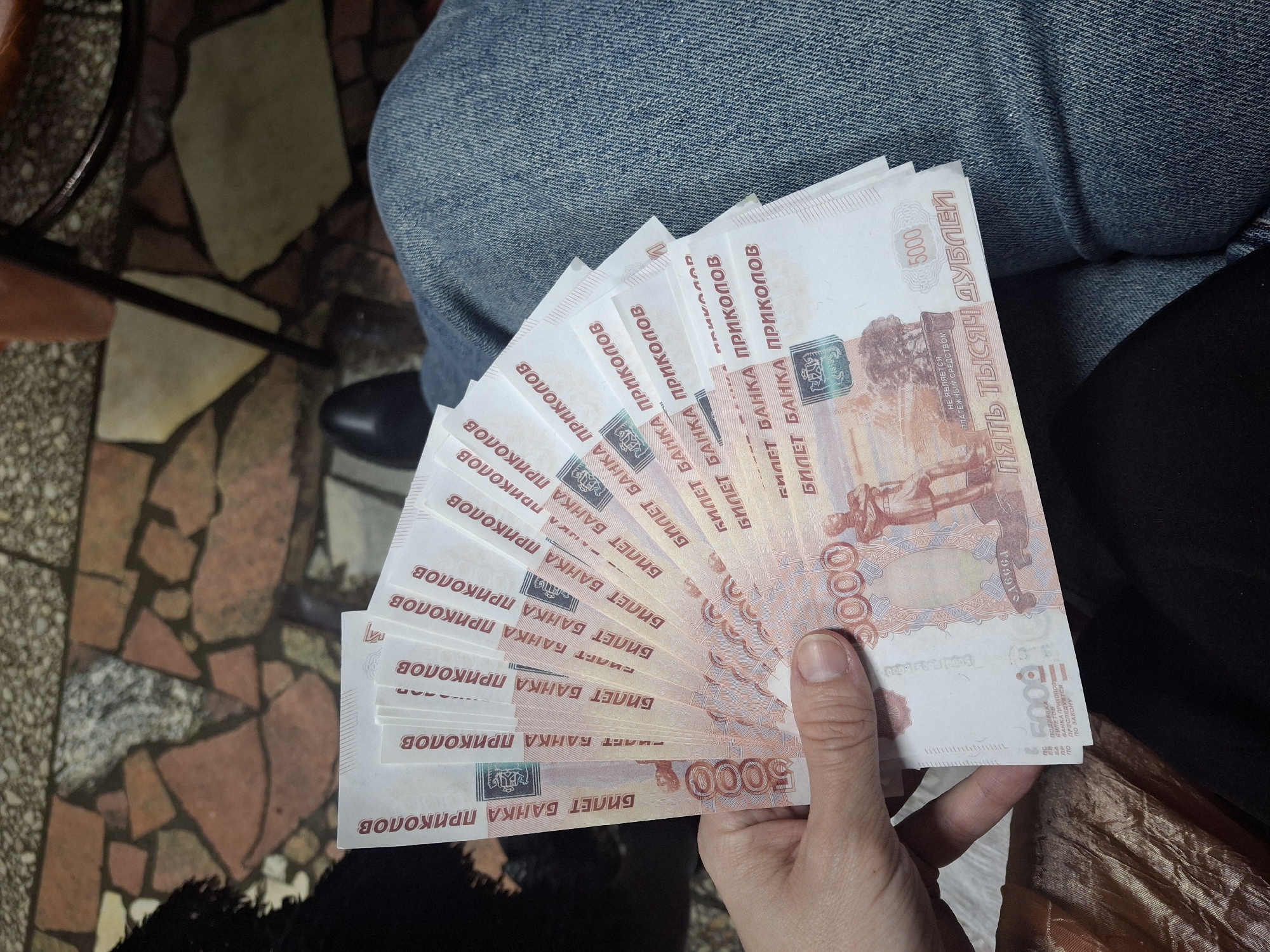 A person holds a fan of ten Russian 5000 "ticket bank pricolov" (joke bank tickets) in their right hand. The tickets are white with red writing and images. The person's hand is light-skinned and the fingernails are trimmed. They are wearing blue jeans. The background includes a mosaic tile floor, a black leather shoe, and an orange-brown wooden chair. The location is not specified as Chaikovsky, Russia.