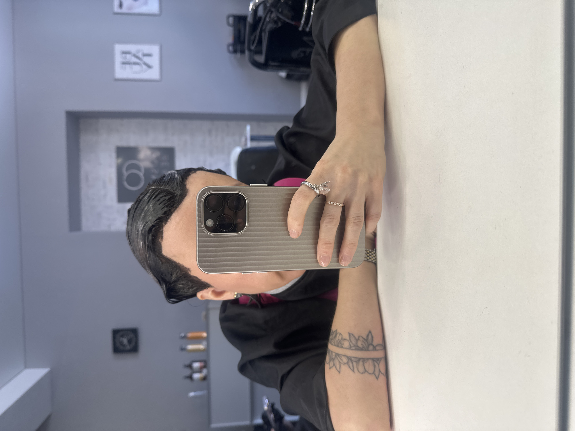 A person, likely female, is taking a mirror selfie in what appears to be a salon or beauty studio in Votkinsk, Russia. The image is taken from a slightly elevated, side-on perspective, with the person's right arm extended to hold a smartphone that obscures most of their face.
The person has dark hair, which is slicked back and appears wet or treated, exposing the scalp around the hairline. They are wearing a black short-sleeved top or smock, with a hint of a magenta or red collar visible underneath. On their right hand, they wear several rings, including a prominent clear stone ring on the ring finger and a thin band with small stones on the middle finger. Their left forearm is visible, featuring an armband tattoo with a floral or leafy pattern. A small, gold-colored earring is visible on the right ear.
The smartphone, held horizontally in a ribbed, metallic-grey case, has a multi-lens camera array visible on its back. The person's right hand is holding the phone, while the left arm rests on the reflective surface, which could be a large mirror or a polished white counter.
The background, seen as a reflection in the mirror/surface, reveals a modern interior with light grey walls. Several framed items are mounted on the wall. One distinct framed sign shows the Chinese character "妆" (Zhuāng), which means "makeup" or "adornment." Below it, another framed item displays a stylized "6" or "b" within a circular logo. Other elements in the background reflection include what appears to be salon equipment or shelves with bottles. The overall setting suggests a professional beauty or hair treatment environment. There are no explicit indicators of time of day or weather, and the person's emotions are not discernible.