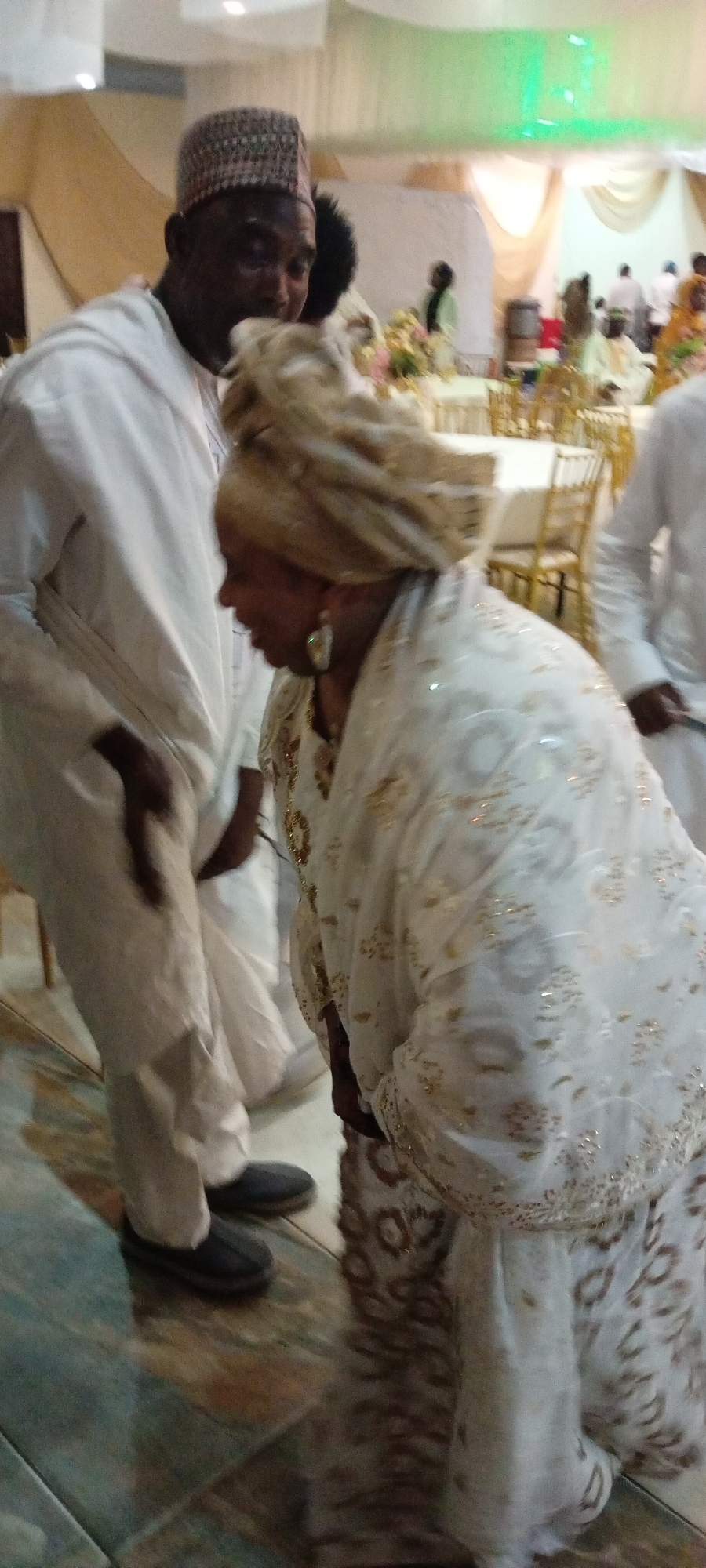 The image captures a cultural gathering, possibly a wedding or naming ceremony, at an indoor venue in Gandu Albasa, Nigeria. Two individuals are prominently featured in the foreground. On the left, a man wearing a white traditional outfit and a patterned cap is partially visible, appearing to lean in towards something out of frame. His attire suggests a formal occasion. To his right, a woman is the central focus, dressed in an elaborate white traditional garment adorned with gold embroidery and beadwork, and a matching headscarf. She is captured in a low bending posture, her face turned downwards, possibly interacting with someone or something below her. Her large earrings add a touch of elegance. The background reveals elements of the venue, including draped fabric hangings, chairs, and tables, indicating a reception or banquet setting. The floor is tiled with a mosaic pattern of green, blue, and brown hues, reflecting the overhead lighting. Several other individuals are visible in the middle and far background, also in traditional attire, suggesting a communal event. The overall ambiance is festive and celebratory, common for such cultural ceremonies in Nigeria.