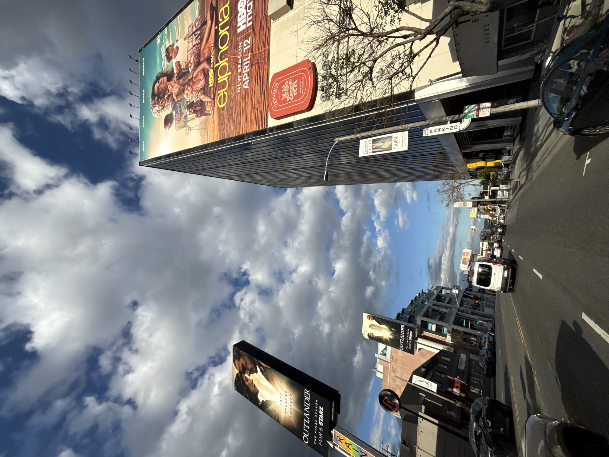 The image captures a street scene in West Hollywood, United States of America, under a sky with dramatic cloud formations. The view is from a street level perspective.
The scene is dominated by billboards advertising TV shows. A large billboard on the side of a tall building showcases "Euphoria" (HBO), advertising its new season with a set date of April 12. Beneath it, a sign for "Christian's Wine Bar & Restaurant" is visible. Further down the street, another billboard advertises the final season of "Outlander" on Starz, with a March 6 release date. A sign reads "RAIN."
Traffic populates the street, with vehicles, including a white van, visible along the asphalt roadway. Buildings line the street, with visible storefronts. A parking sign is visible. Branches of trees are present.