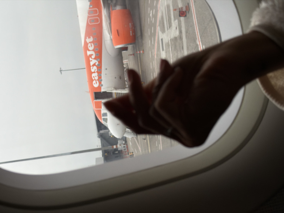 The image captures an exterior view of an airport tarmac from the interior of an aircraft through a passenger window. The scene is located at Lyon-Saint Exupéry Airport (Aéroport de Lyon-Saint Exupéry) in Colombier-Saugnieu, France, under an overcast sky during daylight hours.
In the immediate foreground, a person's right hand and wrist are visible, silhouetted against the brighter exterior. The hand is loosely clasped or resting, with a ring present on the ring finger. The cuff of a light-colored garment is visible around the wrist.
Beyond the window, a large orange and white easyJet Airbus A320 series aircraft is parked on the tarmac. The front section of the aircraft, including its nose, cockpit windows, and the main passenger door (L1), is visible. The passenger door is open, and a set of mobile boarding stairs is positioned at the entrance. The "easyJet" logo is prominently displayed in orange text on the aircraft's white fuselage. The right engine, with its distinctive orange cowling, is also visible. The tarmac features various white and red ground markings, and several orange traffic cones are positioned around the easyJet aircraft. In the background, to the left of the easyJet aircraft's nose, a grey jet bridge structure is discernible, connecting to another terminal or gate. A tall, multi-lamp light pole stands in the mid-background.
