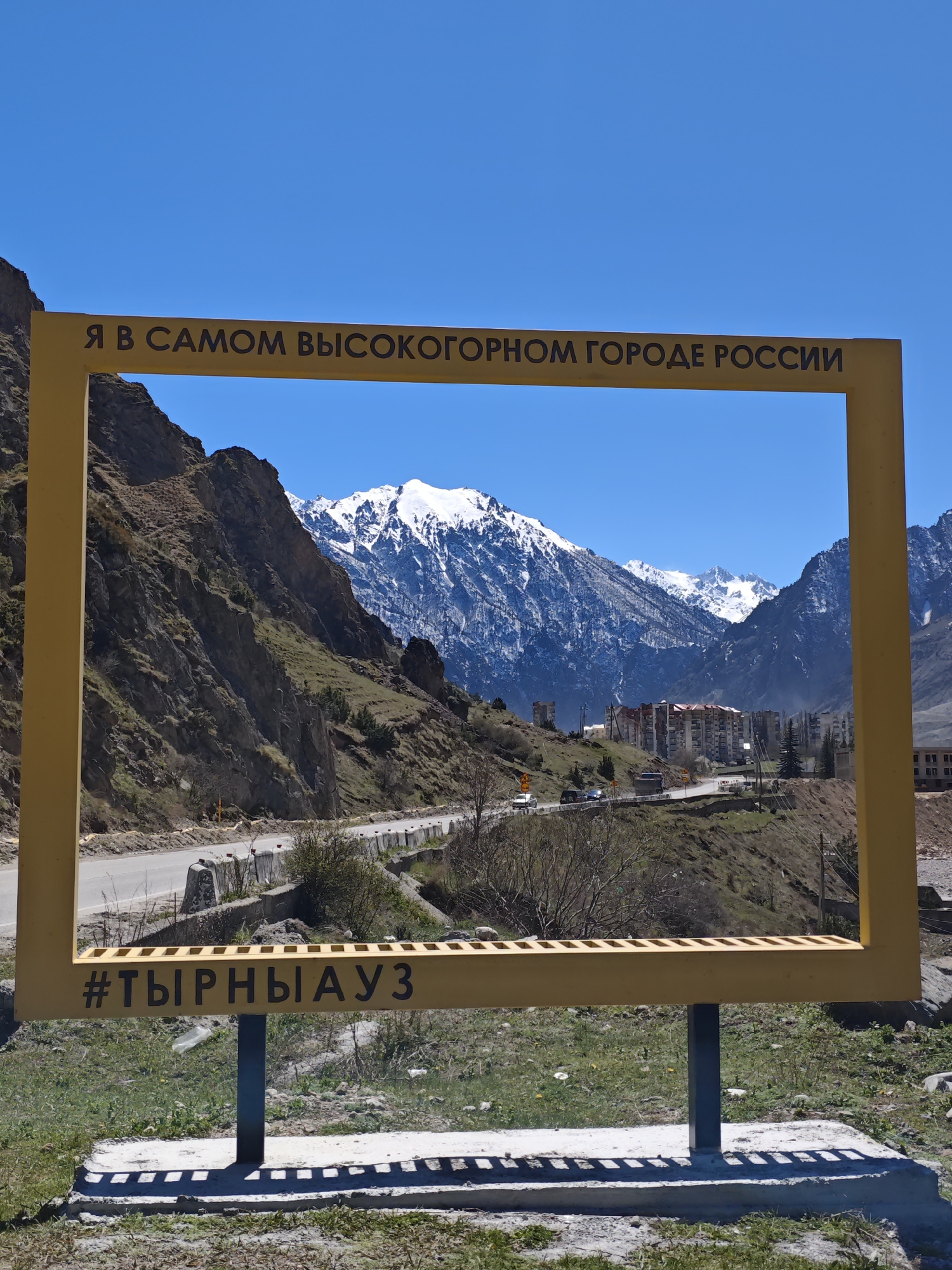 The image depicts a yellow frame structure positioned in Tyrnyauz, Russia, against a backdrop of mountainous terrain. The frame bears the inscription "Я в самом высокогорном городе России" (I am in the highest mountain city in Russia) on its top section and "#ТЫРНЫАУЗ" (#TYRNYAUZ) on its bottom section. The backdrop showcases a landscape featuring a road winding along a mountainside, buildings nestled amidst the hills, and snow-capped peaks in the distance under a clear blue sky. The foreground shows the grassy area where the frame stands, casting shadows on the ground. The scene is bathed in sunlight, highlighting the contrast between the yellow frame and the natural surroundings.