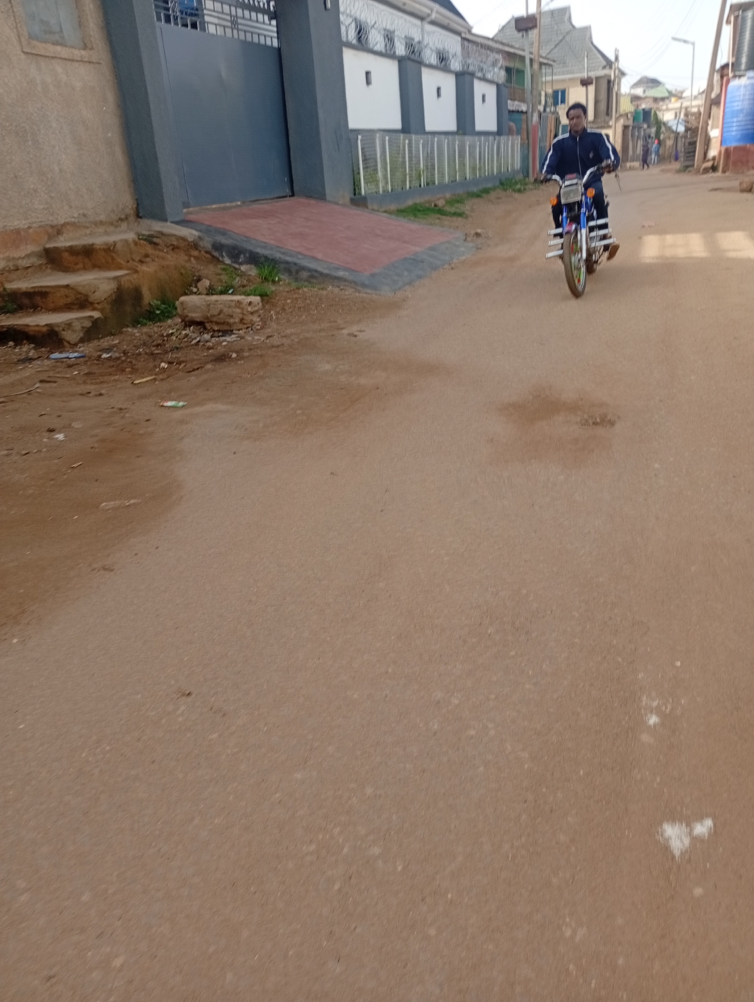 A man rides a motorcycle down a dirt road in a residential area. The man is wearing a dark blue jacket with white stripes on the sleeves. The motorcycle is blue and has a metal rack on the front. In the background, there are houses and a large gate. The road is dusty and uneven. The scene appears to be in Rafin Par, Nigeria.