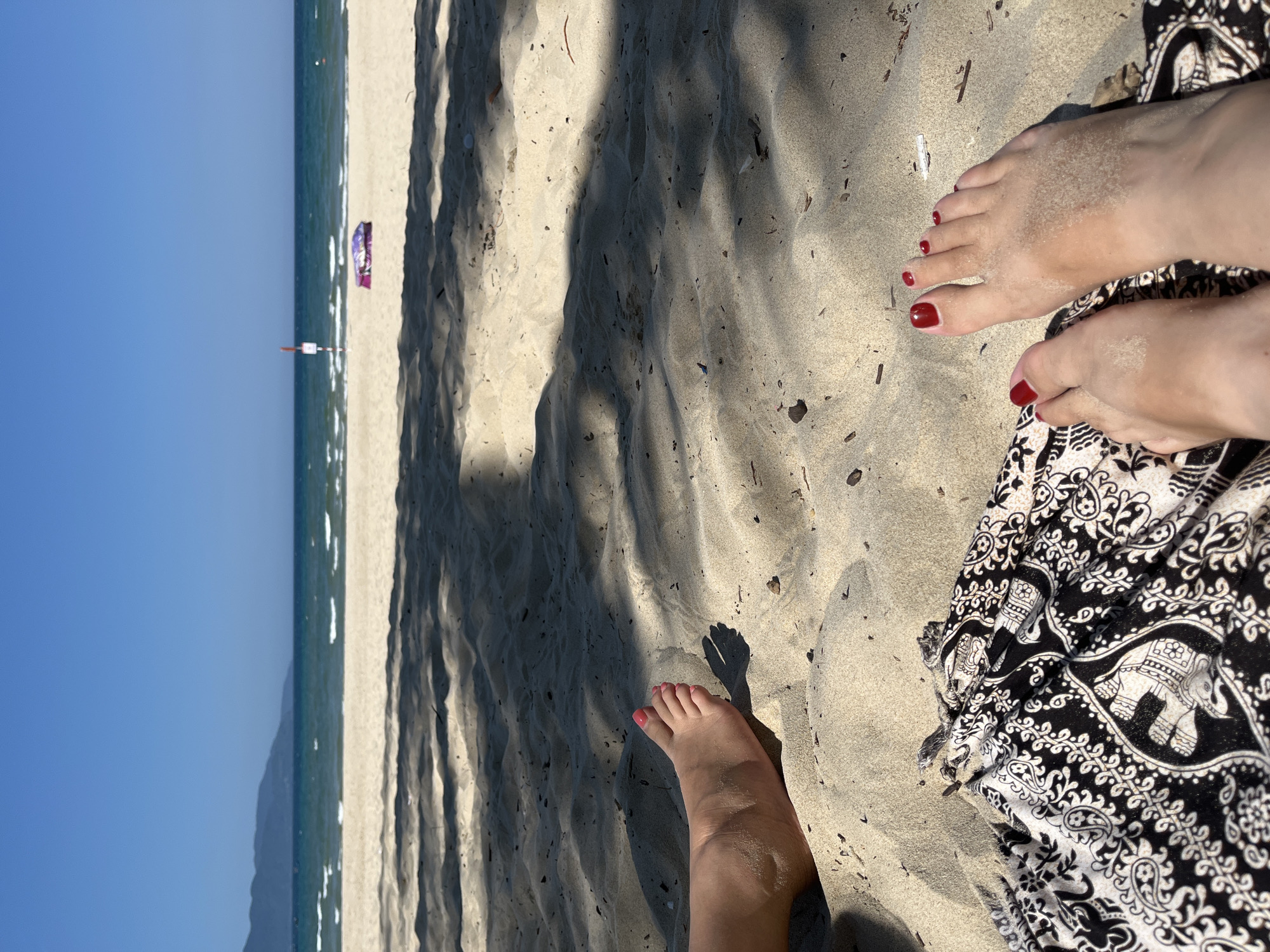 The image shows a view of a beach in Đà Nẵng, Vietnam, from a ground-level perspective. The scene includes sand, the ocean, and a clear blue sky. In the foreground, a person's feet are visible, displaying red nail polish. The feet are partially covered in sand and are positioned near a black and white patterned garment. In the distance, a beach umbrella and a pole are seen along the shoreline. Shadows from trees or other objects are cast across the sand.