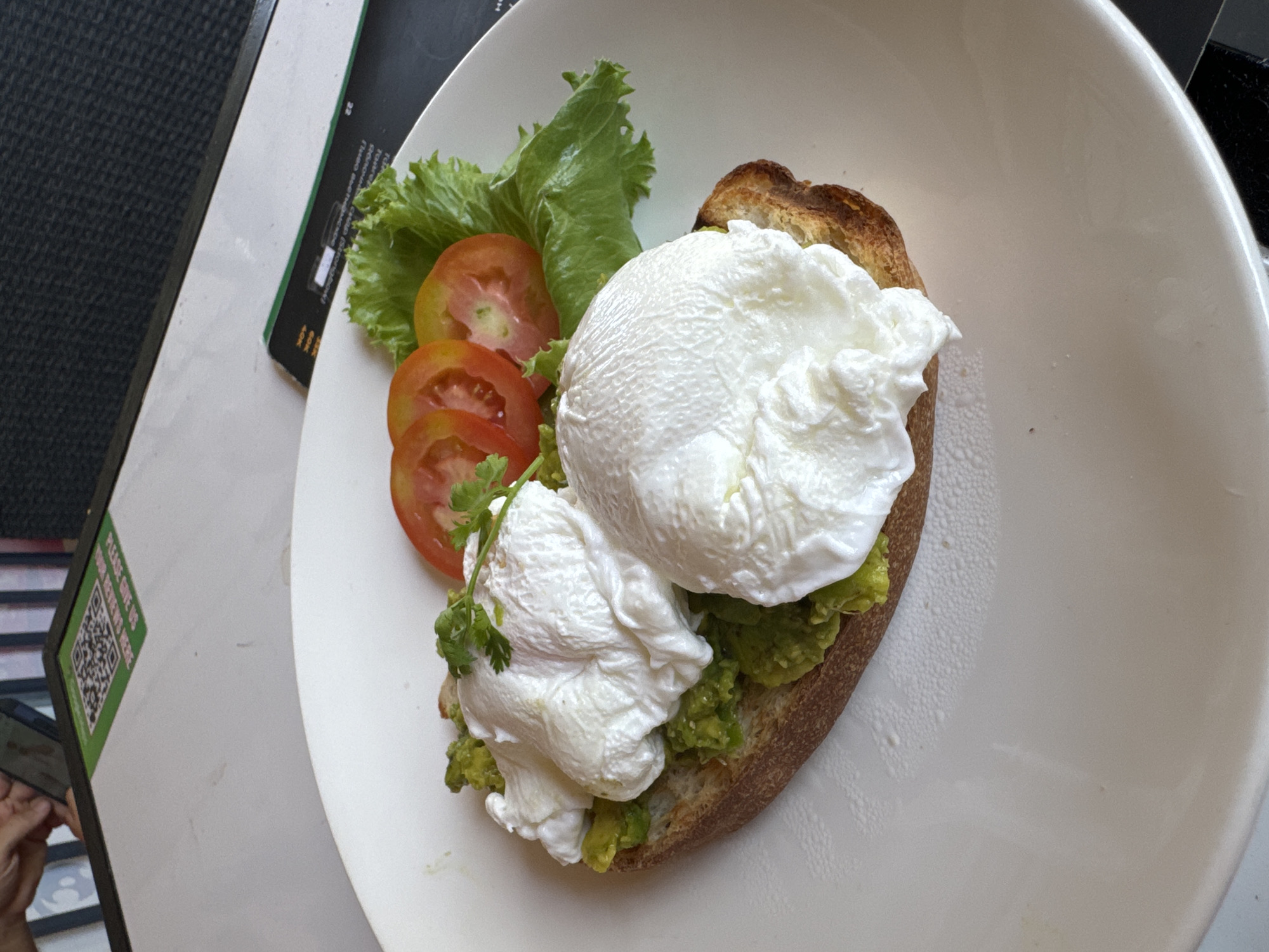 The image shows a plate of food, likely in a restaurant in Mũi Né, Vietnam. The dish features two poached eggs on toast spread with mashed avocado. The plate also includes several slices of tomato and a piece of lettuce. The plate is white and sits on a table, with a menu and a QR code for leaving reviews visible.