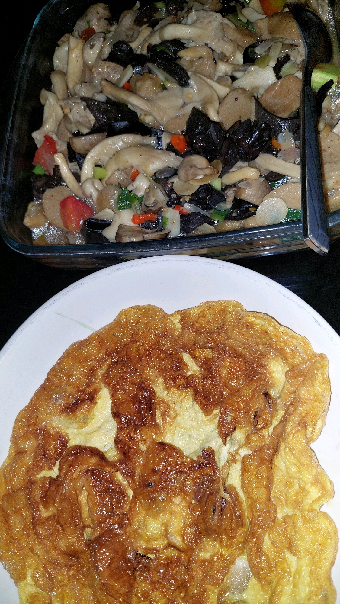 An overhead shot shows a meal that includes an omelet on a white plate and a glass dish containing a mushroom and vegetable stir-fry. The omelet is golden brown and appears to be fluffy. The mushroom and vegetable stir-fry is a mix of light-colored mushrooms, dark mushrooms, diced red tomatoes, and chopped green onions. The meal is presented on a dark surface, likely a table. No specific location in Bekasi, Indonesia, is visible.