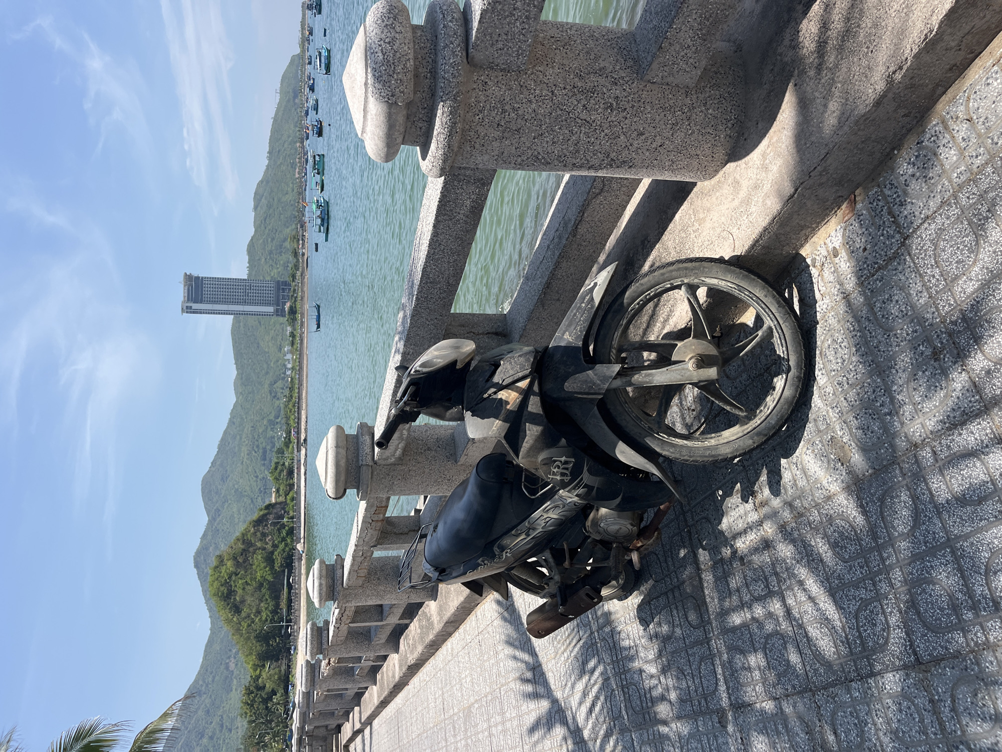 The image depicts a sunlit coastal scene in Bac Nha Trang Ward, Vietnam. In the immediate foreground, a weathered black underbone motorcycle is parked on a grey, patterned tiled promenade. Strong shadows, including those from palm fronds and the motorcycle itself, fall across the pavement. A light grey concrete balustrade with decorative cylindrical posts separates the promenade from the tranquil, light blue-green sea. Approximately 20-30 small, brightly colored wooden fishing boats, predominantly blue and green, are anchored in the calm waters of the bay. The midground features a curving coastline with lush green, tree-covered hills and a sandy stretch. In the background, a tall, modern high-rise building, appearing to be a hotel or residential tower, stands prominently against a backdrop of distant green mountains. The sky is clear and light blue with scattered wispy white clouds. No individuals are visible in the scene.