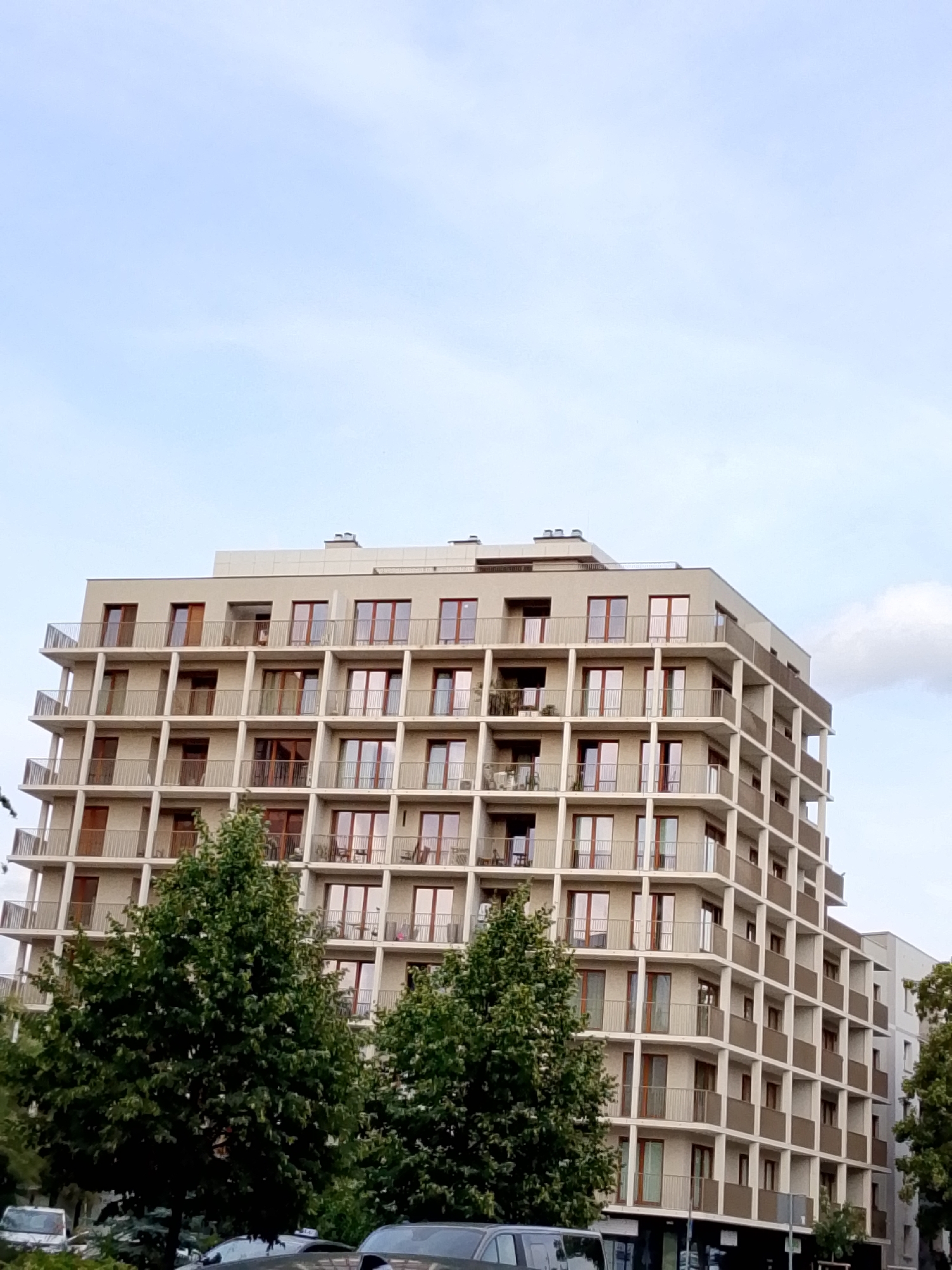 The image captures a modern, multi-story residential building located in Wrocław, Poland. The building features a facade composed of repeating square and rectangular balconies and windows, creating a grid-like pattern. The balconies are finished with light-colored railings, and the windows appear to have dark frames and wooden doors. Trees are visible in the foreground, partially obscuring the lower levels of the building and the street. Several cars are parked or driving on the street in front of the trees. The sky above is a pale blue with some scattered clouds. The overall scene depicts an urban residential environment.