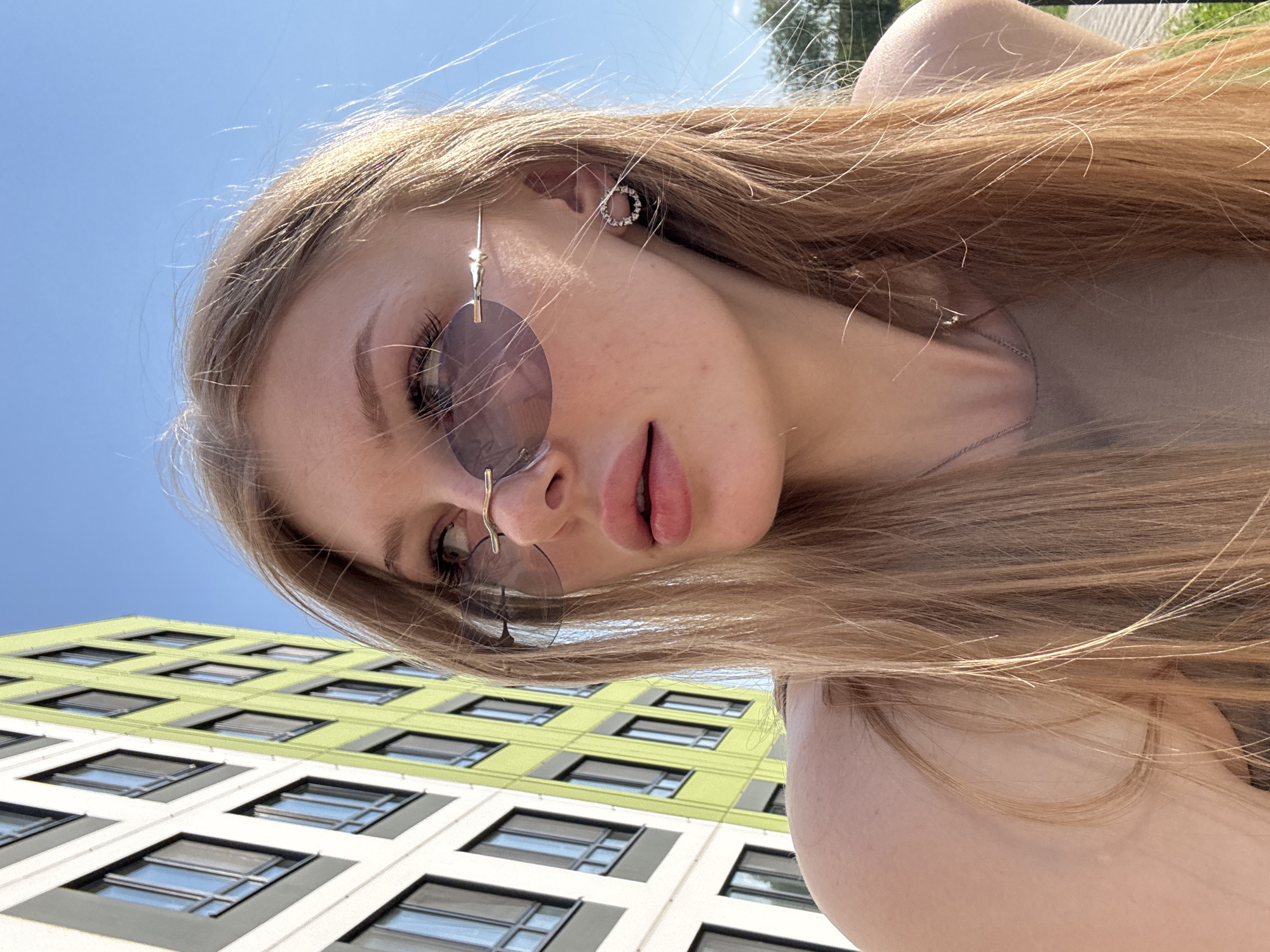 A photograph taken from a low angle features a blonde woman with light skin, wearing round sunglasses with a unique nose bridge design and circular earrings. She appears to be outdoors, with a modern building partially visible in the background, featuring a grid of square windows and alternating green and white colored walls. Above the building, the sky is clear and blue. The location is identified as Perm, Russia. The woman is looking down, and the composition suggests she is taking a selfie.