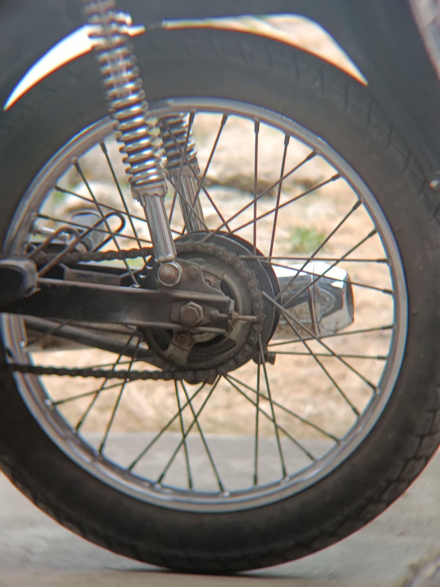 This is a close-up, eye-level shot of a motorcycle's rear wheel and suspension system. The image is focused on the spokes, chain, sprocket, and shock absorbers. The tire is black and appears to have some wear. The background is blurred and suggests an outdoor environment with dirt or gravel. The overall impression is of a functional, perhaps older, motorcycle. The location is identified as Sarwadadi, Indonesia.