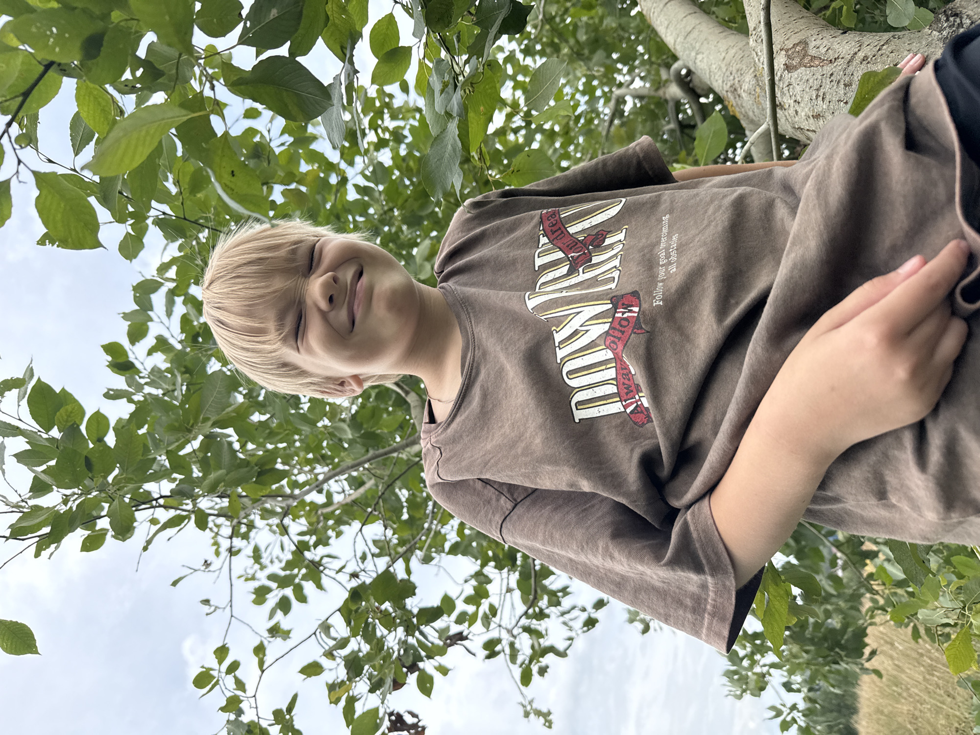 A young person with blonde hair and a brown t-shirt is in a tree. The t-shirt has text and graphics on it. The person is smiling with their eyes closed. Leaves and branches of a tree are in the foreground and background, with a cloudy sky visible. The overall tone is casual and relaxed, possibly indicating a warm day. The image appears to be taken outdoors in a natural setting, possibly a park or backyard. The text on the t-shirt reads "DREAM ON" and "Follow your goal overcoming all obstacles."