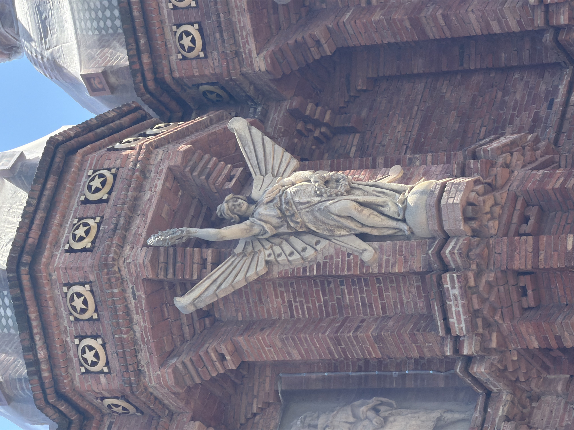 The image shows a close-up, low-angle shot of the Arc de Triomf in Barcelona, Spain. The monument, constructed from red brick, features an angelic statue with outstretched wings holding a wreath in its left hand and a bundle of branches in its right. The statue is positioned on a pedestal against the brickwork of the arch. Decorative elements, including square panels with stars, are visible on the brick facade. The sky is clear and blue.