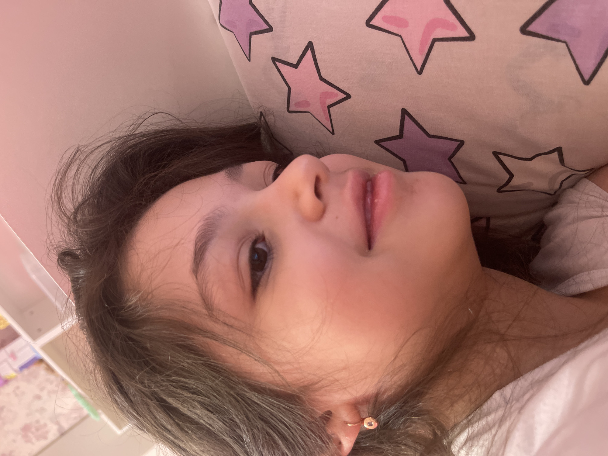 A close-up, slightly rotated portrait shot of a young girl. She is lying on a pillow with a star pattern. Her face is angled towards the camera, showing her right eye, nose, and mouth. She has brown hair and an earring in her left ear. The background includes a white shelf. The location cannot be determined with certainty to be Yessentuki, Russia, based solely on this image.