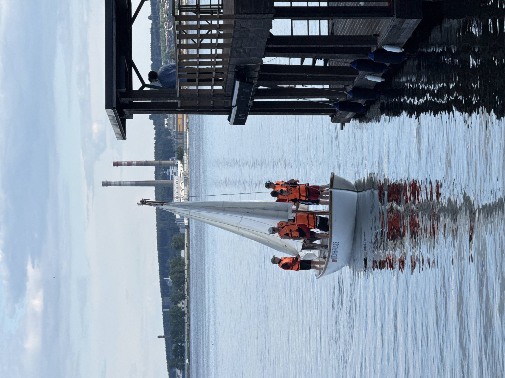 This daytime image, captured in Votkinsk, Russia, presents a scene on a body of water, likely a lake or wide river, adjacent to an industrial shoreline. In the foreground-midground, a small white sailboat with a single mast and a furled or partially furled white sail is navigating calm water. Six individuals, all wearing bright orange life vests, are standing on the sailboat's deck. The boat bears the registration number "VBO056RUS18" on its hull. To the right, a two-story, dark wooden pier or structure extends from an unseen shore, with its lower section partially submerged in the water. Several dark blue and white fenders are suspended from the side of the pier. On the upper level of this structure, one person stands with their back to the viewer, facing out towards the water. The background features a distant tree-lined shore. Prominently positioned on the horizon are several industrial facilities, including two tall, red-and-white striped smokestacks and other grey industrial buildings. The sky above is partly cloudy with patches of blue.