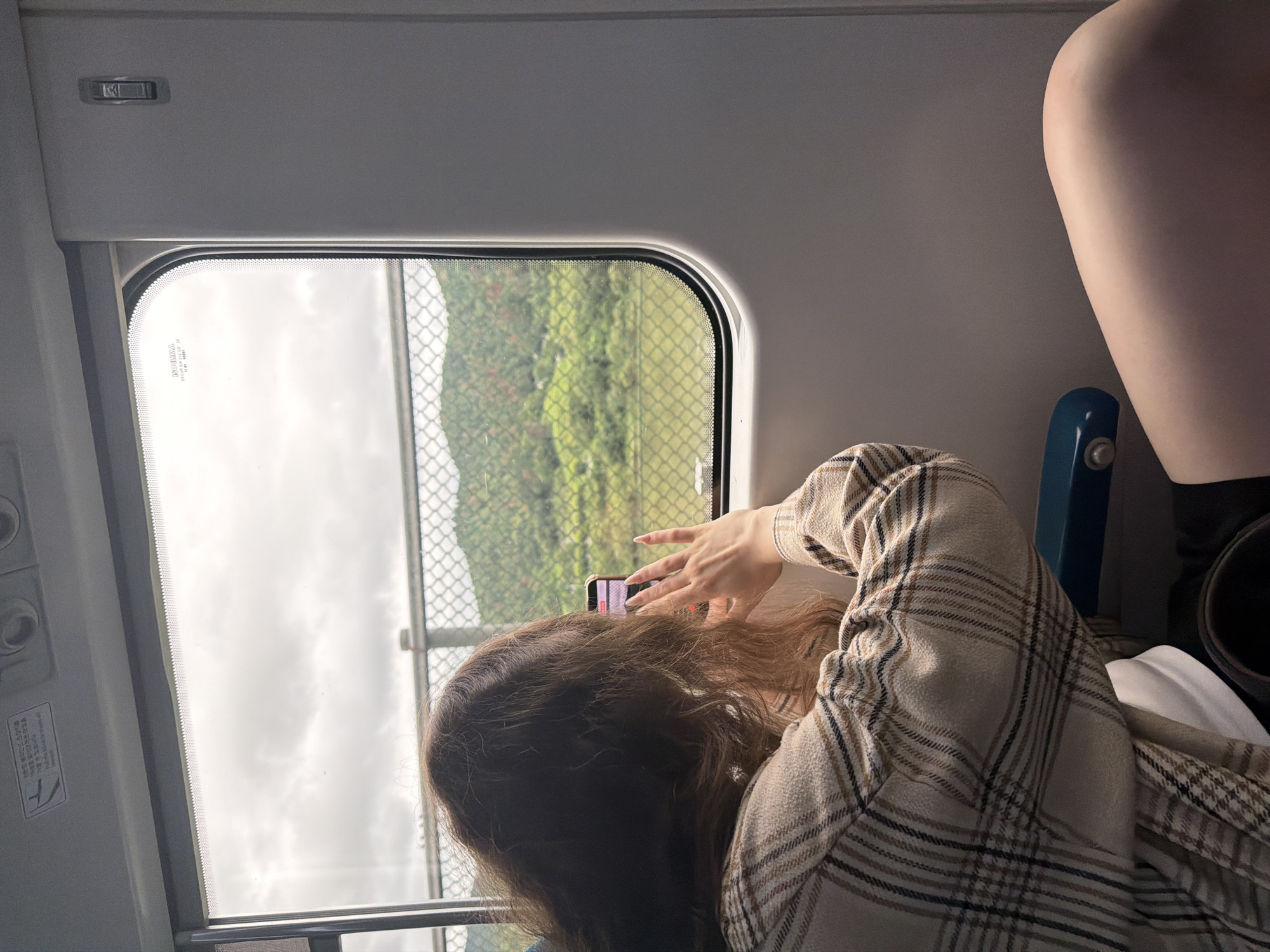 A young woman with long, brown, wavy hair, wearing a plaid jacket over a white top, is seated by a train window. She is holding a smartphone in her right hand, seemingly taking a picture or video of the passing scenery. Her left hand is near the phone, with manicured fingernails visible. Outside the train window, a green, mountainous landscape is visible under a cloudy sky. A wire fence is positioned between the train and the landscape. The interior of the train car is visible, including a seat armrest and the gray wall of the train. The image appears to have been taken in Chuncheon-si, South Korea, possibly from a tourist train or intercity service.