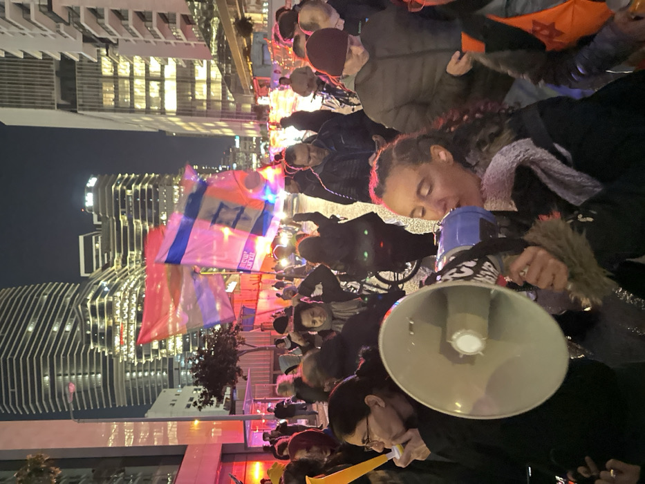 A crowd gathers at night, illuminated by city lights. Prominent in the foreground, a woman speaks into a megaphone. People wave Israeli flags adorned with lights. Buildings rise in the background. The atmosphere suggests a protest or demonstration.