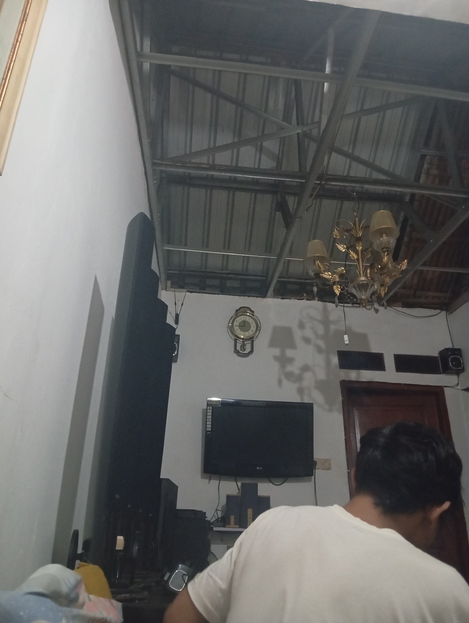 The image depicts an interior scene, possibly a living room or bedroom, with a person seen from the back wearing a white t-shirt. The room has a high ceiling with exposed metal beams and corrugated metal roofing. A chandelier hangs from the ceiling. On the wall, there is a round analog clock and a television set. To the left of the television, a tall, dark, rectangular object stands vertically, possibly a speaker or a piece of furniture. The lighting appears dim, with shadows cast on the wall from the chandelier. The overall impression is of a functional but somewhat unfinished or industrial living space.