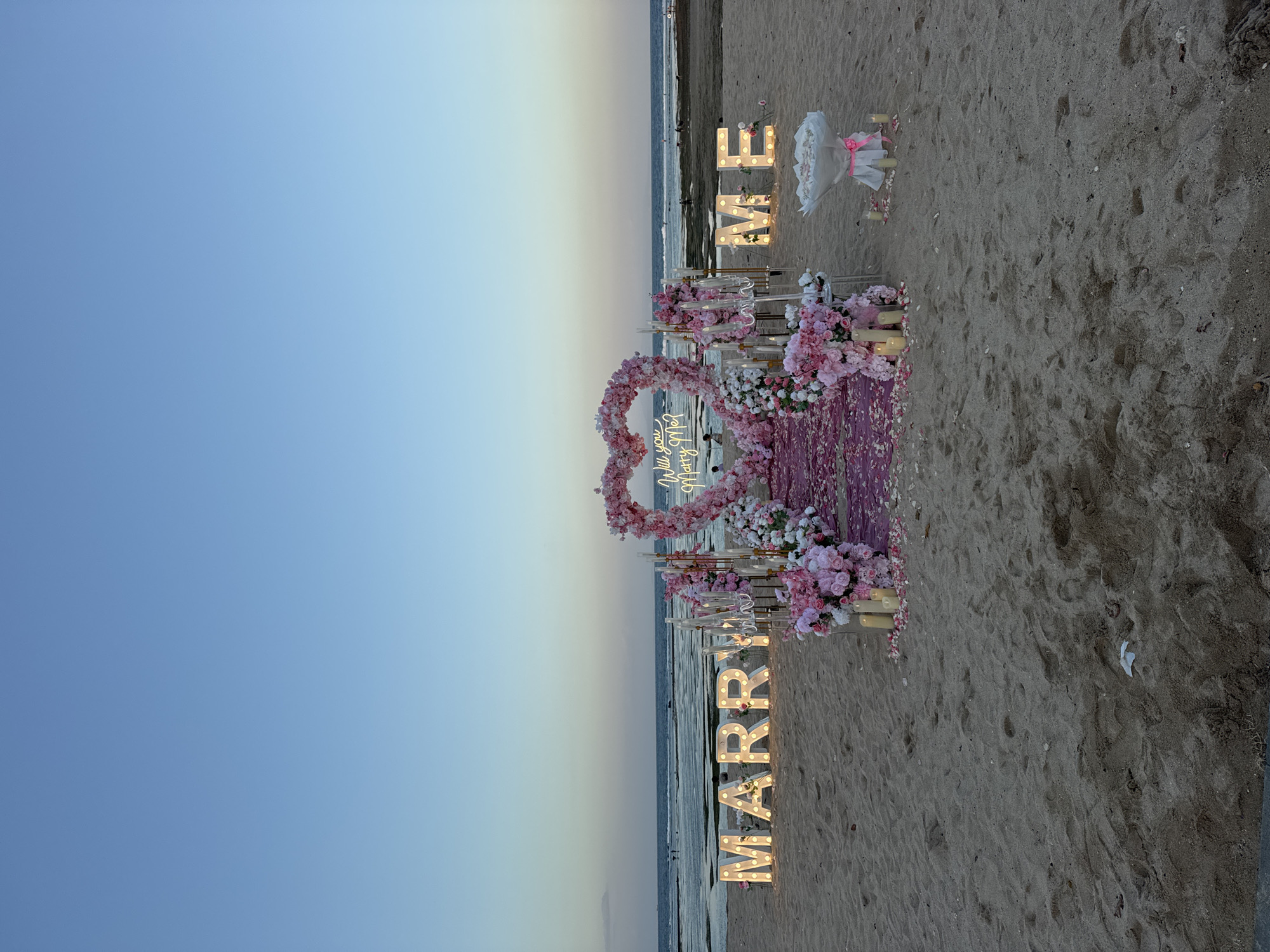 A proposal setup is displayed on a sandy beach at dusk, likely in Kuta Selatan, Indonesia, given the tropical setting. The scene is dominated by decorative elements arranged on the sand facing the ocean.
In the center, a large heart-shaped arch adorned with pink and white flowers frames a neon sign that reads "Will you Marry Me?". In front of the arch, a pink carpeted aisle is scattered with flower petals. Large, illuminated block letters spelling "MARRY" are positioned to the left of the arch, and "ME" to the right. These letters are also decorated with small pink flowers.
Flanking the aisle are decorative arrangements of pink and white flowers and lit candles. To the right of the "ME" letters, a bouquet of flowers wrapped in white paper and tied with a pink ribbon sits on the sand, also surrounded by a few candles.
The ocean is visible in the background, with gentle waves lapping at the shore. A few people are visible in the distance near the water, and some are in the water itself. The sky is a clear gradient of blue, transitioning from a lighter hue near the horizon to a deeper blue above. The overall ambiance is romantic and celebratory, suggesting a planned marriage proposal. The sand is textured with footprints.