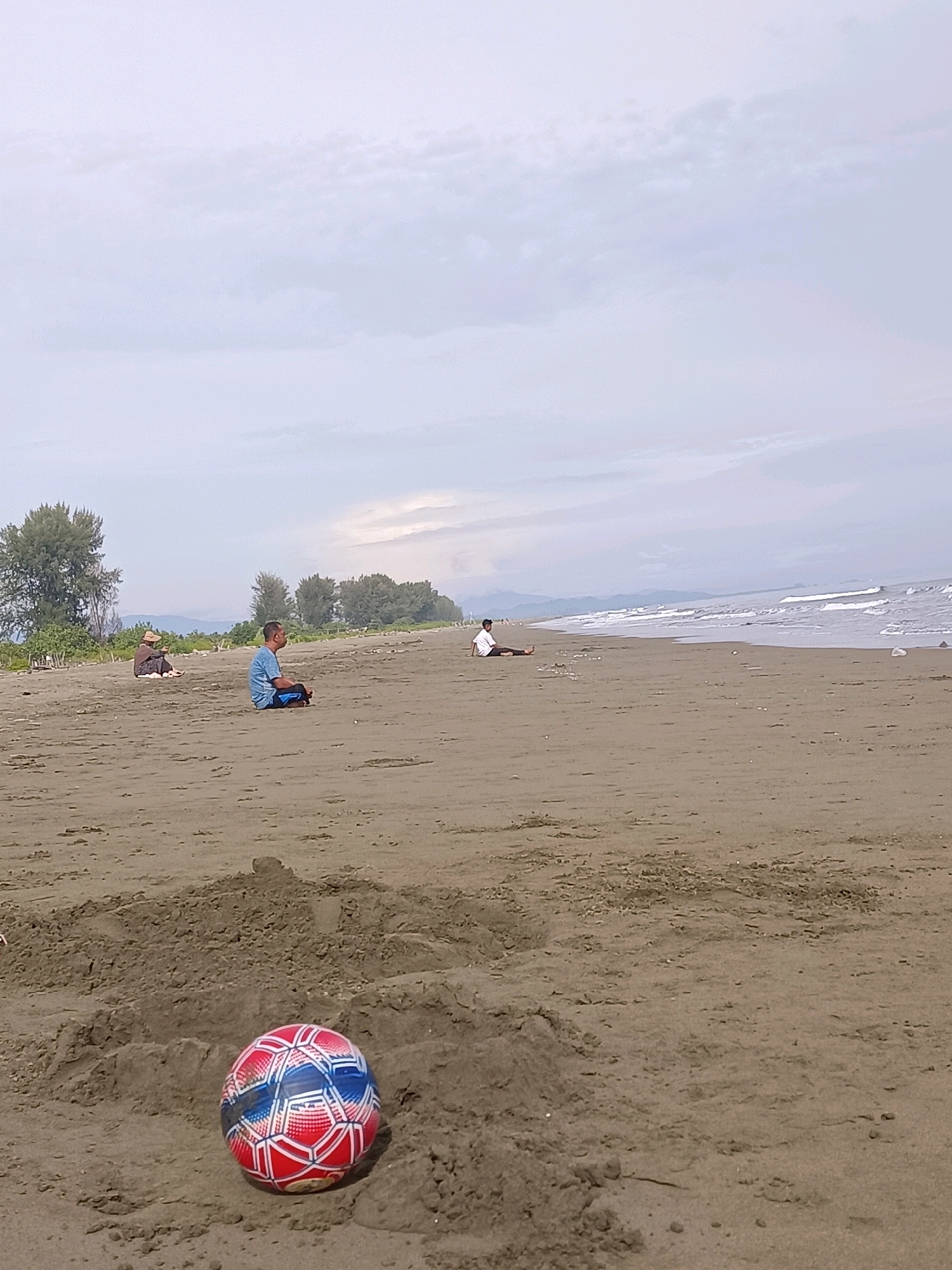 A wide, sandy beach in Reudeuep, Indonesia, is depicted under an overcast sky. In the foreground, a multi-colored soccer ball, predominantly red and blue with white accents, rests partially buried in a mound of sand. Further back, three individuals are scattered across the beach. One person, wearing a straw hat and a dark garment, sits on the sand near a clump of trees. Another person in a blue shirt sits cross-legged. A third person, in a white shirt, lies down near the water's edge. The ocean stretches across the background with gentle waves breaking on the shore. Trees line the far edge of the beach, and mountains are visible in the distant haze. The overall scene conveys a sense of quiet relaxation on a cloudy day.