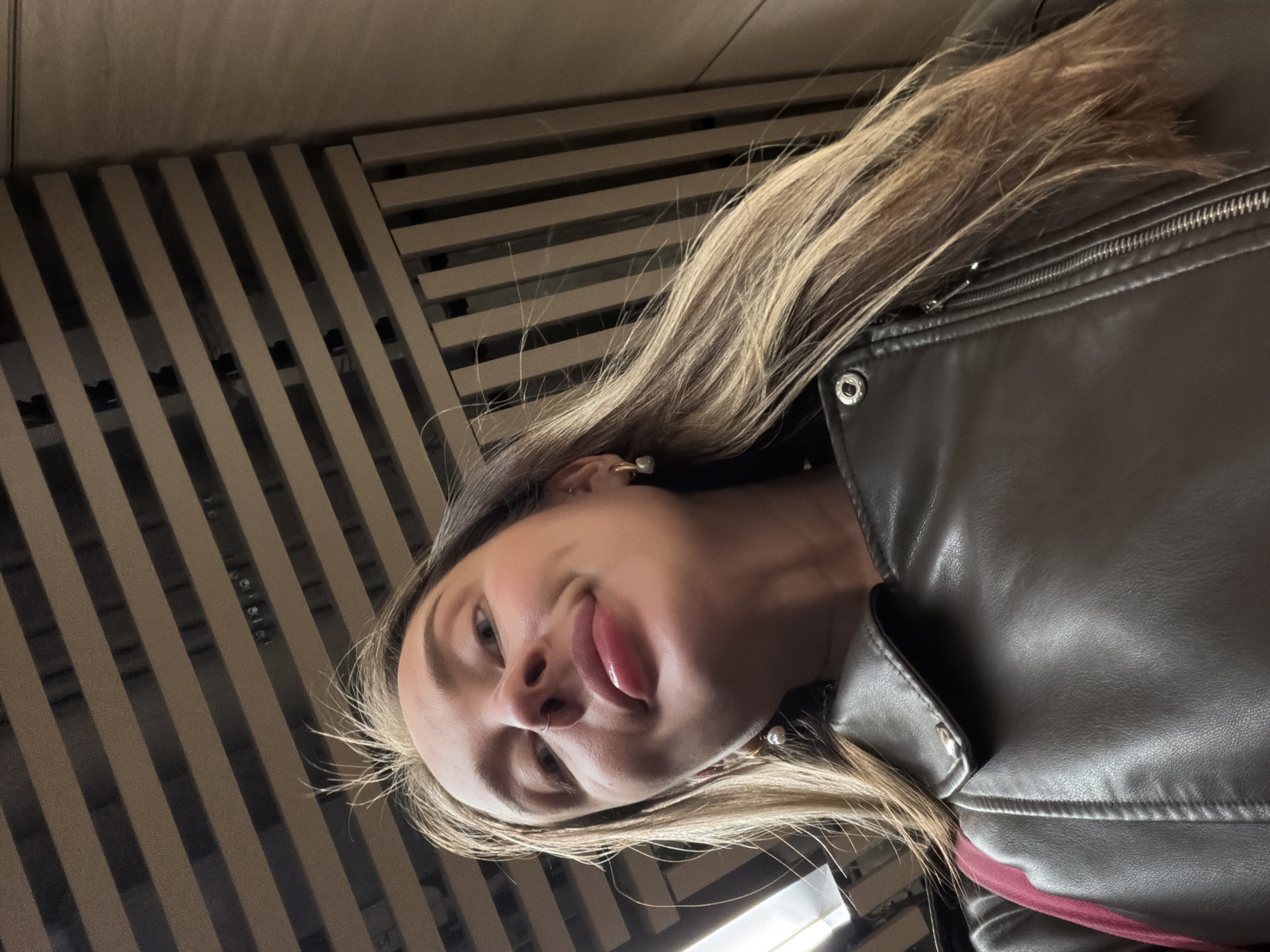 A low-angle portrait of a Caucasian woman with blonde hair, wearing a dark leather jacket. She is sticking her tongue out. The background is a series of horizontal, light-colored slats. The location is unknown and cannot be determined from the image.
