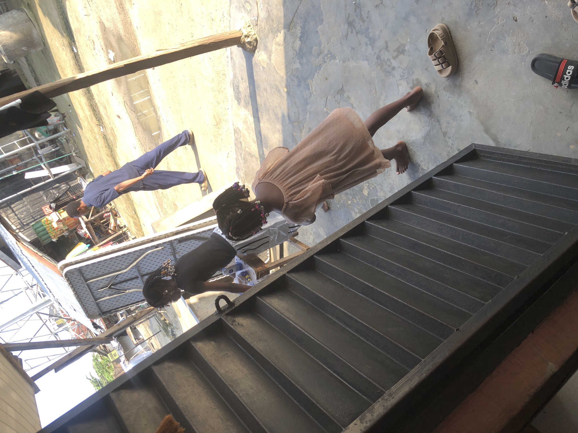 In Ado, Nigeria, a bright, sunlit scene unfolds in what appears to be a market or roadside area. In the foreground, a person is descending a dark, metallic staircase. To the right of the staircase, a young girl in a light-colored dress with ruffled sleeves stands facing away from the viewer, her back to the camera, with her hair in braids adorned with beads. Nearby, a pair of tan sandals rests on the concrete ground. Further in the background, a man in blue pants and a dark shirt walks away from the camera, his left hand extended behind him. Behind him, various structures suggest a commercial or residential setting, including corrugated metal roofs, wooden stalls, and possibly some stacked containers. The ground is a mixture of concrete and dirt, with some puddles of water visible. The overall impression is one of everyday activity in a bustling urban environment.