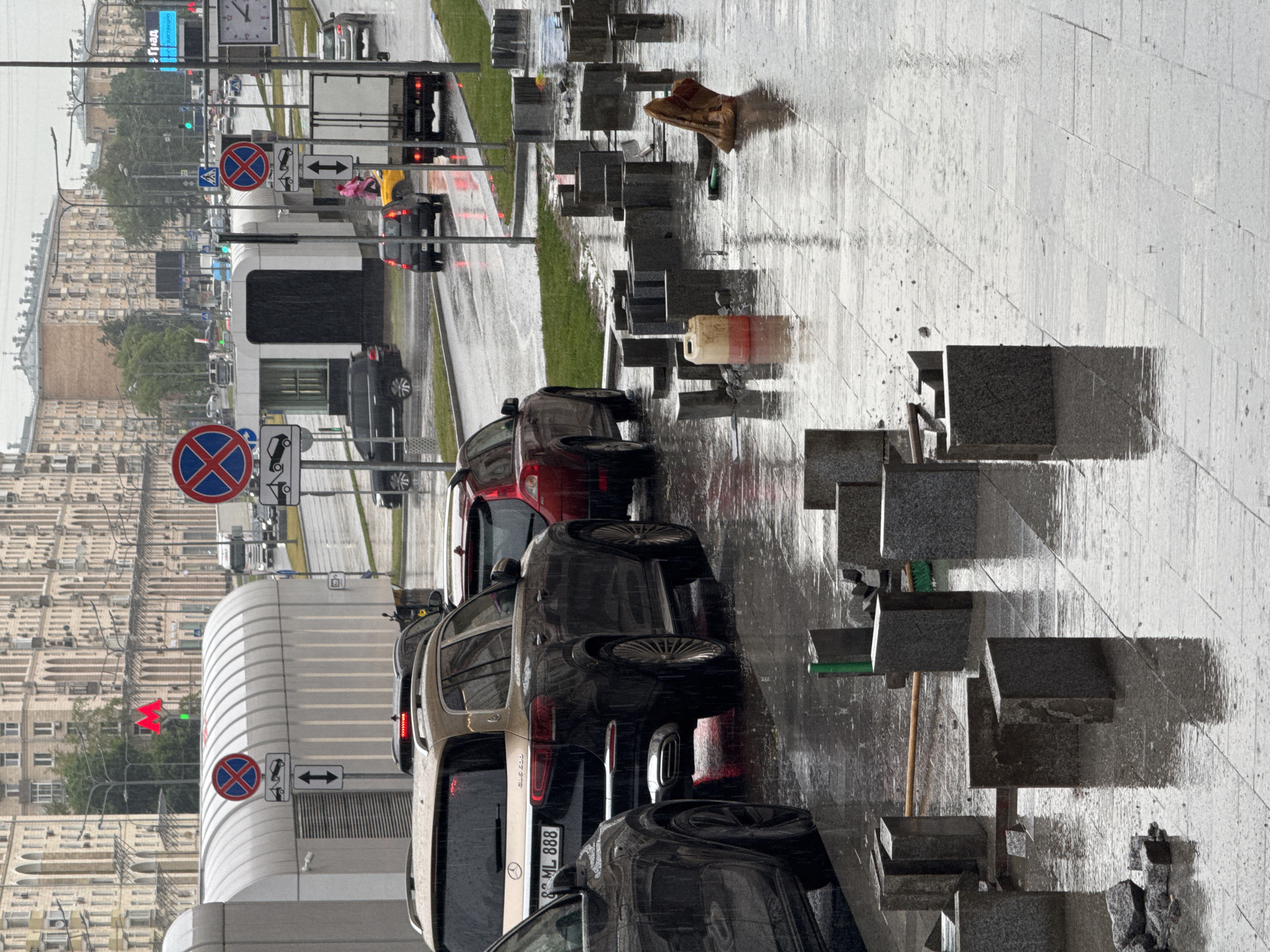 On a rainy day in an unknown city, a street scene unfolds with several vehicles and construction materials scattered across a wet sidewalk. In the foreground, a dark SUV is parked, partially visible on the left. To its right, a luxury SUV, a Mercedes-Benz GLS 600, is prominent, with its license plate reading "ML 888". Further down the street, other cars, including a red Volvo, are seen.
The sidewalk in the foreground is covered with numerous stone blocks, some upright and others lying flat, indicating ongoing or recently interrupted construction work. A yellow plastic container with red liquid, a broom, and a brown paper bag are also present on the wet pavement. Traffic signs, including "No stopping" signs with towing symbols, are visible along the roadside. Buildings with arched windows line the background, suggesting an urban environment. The weather is overcast, and the rain has created puddles and reflections on the street and sidewalk.