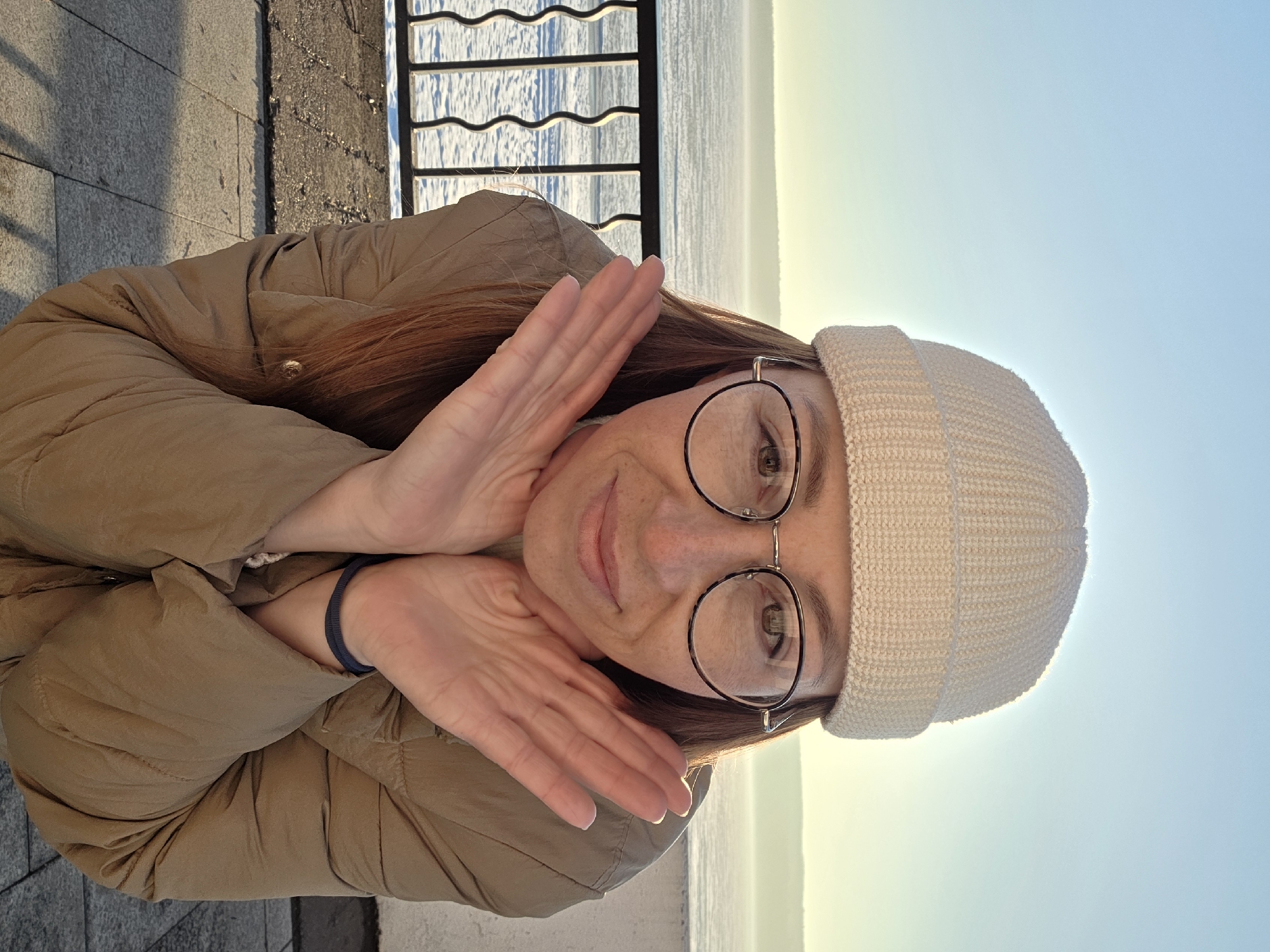 A woman with brown hair and glasses is posing outdoors. She is wearing a tan puffer jacket and a cream-colored knit beanie. Her hands are raised, framing her face. In the background, there is a body of water and a metal fence. The location is Chaikovsky, Russia. The background shows a clear blue sky. The woman appears to be smiling slightly. The setting looks like a sunny day near a waterfront.
