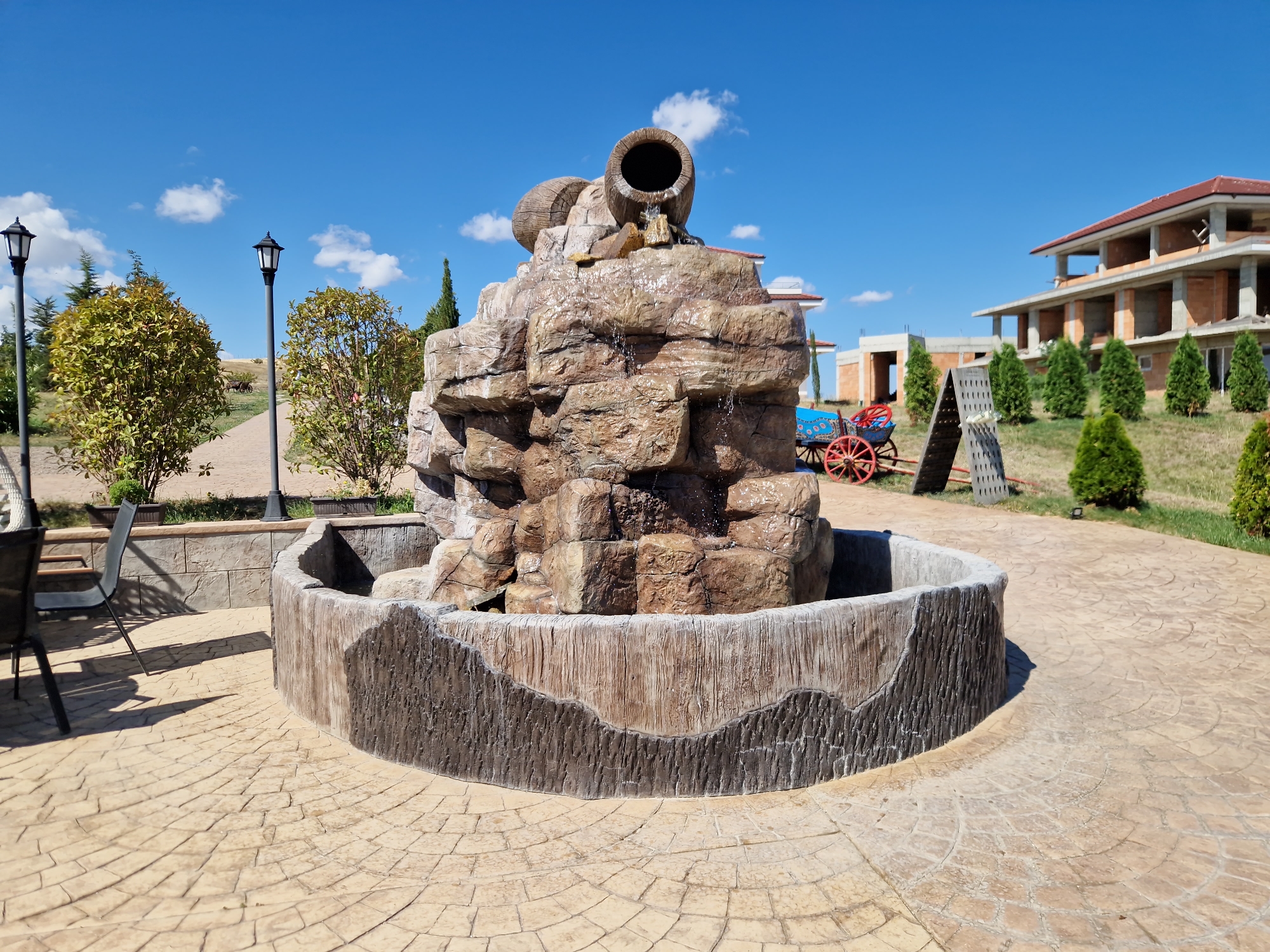 The image depicts an outdoor scene in Tankovo, Bulgaria, featuring a decorative rock fountain with two ceramic barrels at the top, from which water is flowing. The fountain is situated within a curved, textured concrete basin. The surrounding area is paved with stamped concrete in a fan pattern. To the left, there are outdoor chairs and tables. In the background, there are lampposts, various trees and bushes, a partially constructed building, and a small, decorative horse-drawn cart with red wheels. The sky is clear blue with scattered white clouds.