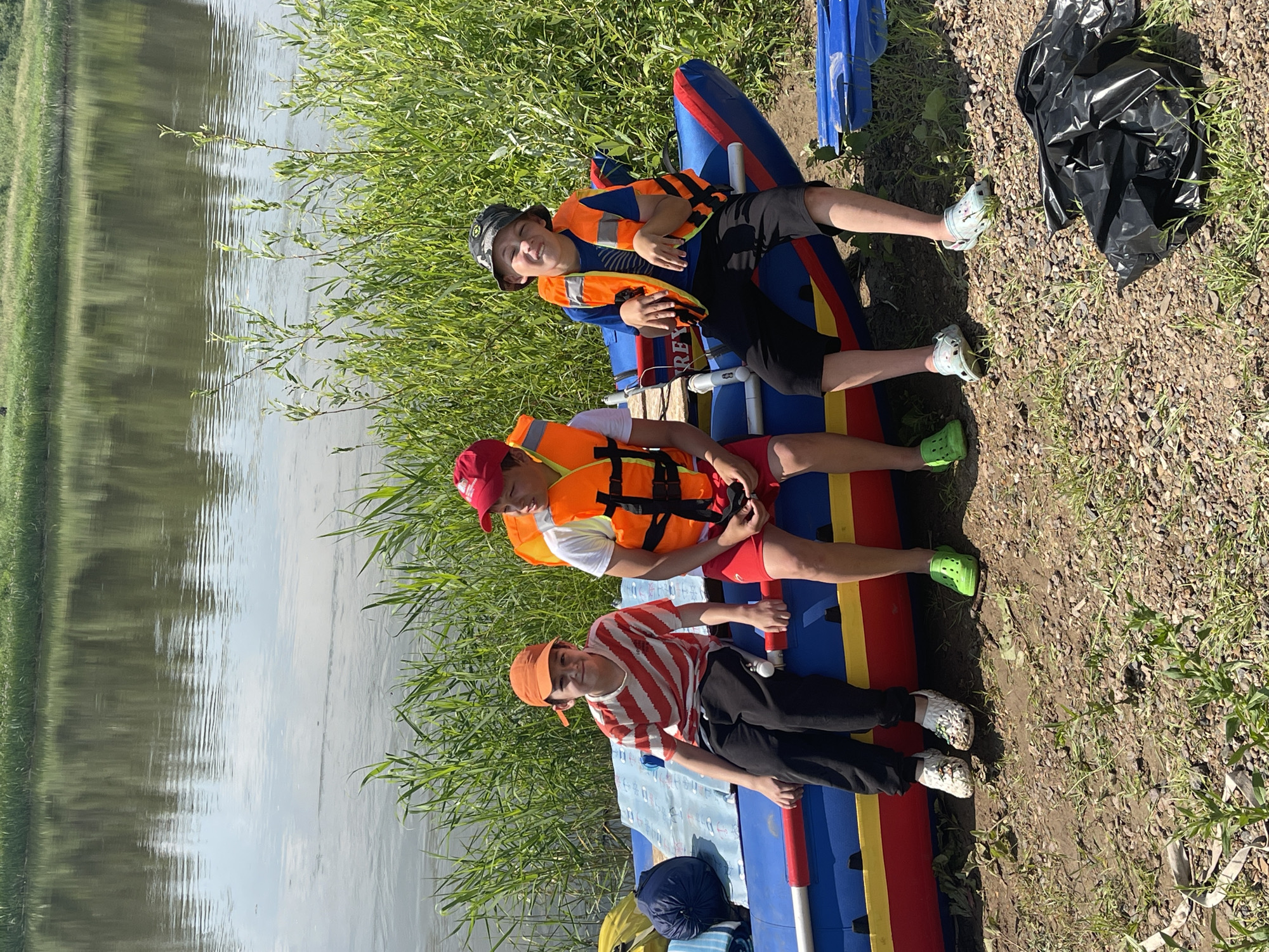 Three young boys, wearing life jackets and casual clothing, are sitting on the edge of a deflated blue and red raft on the muddy bank of a wide, calm river. The boy on the left is wearing an orange cap and a striped t-shirt. The middle boy is wearing a red cap and orange life jacket over a white t-shirt and red shorts. The boy on the right is wearing a camouflage bucket hat and an orange life jacket over a blue t-shirt and black shorts. The river reflects the green foliage on the opposite bank. Tall green grass grows along the bank behind the raft. A dark blue rolled-up sleeping bag and yellow bag are on the raft to the left. A crumpled black plastic bag is on the ground to the right of the raft. The scene appears to be in a natural setting, likely a park or recreational area, near the village of Nezhinka, Russia.