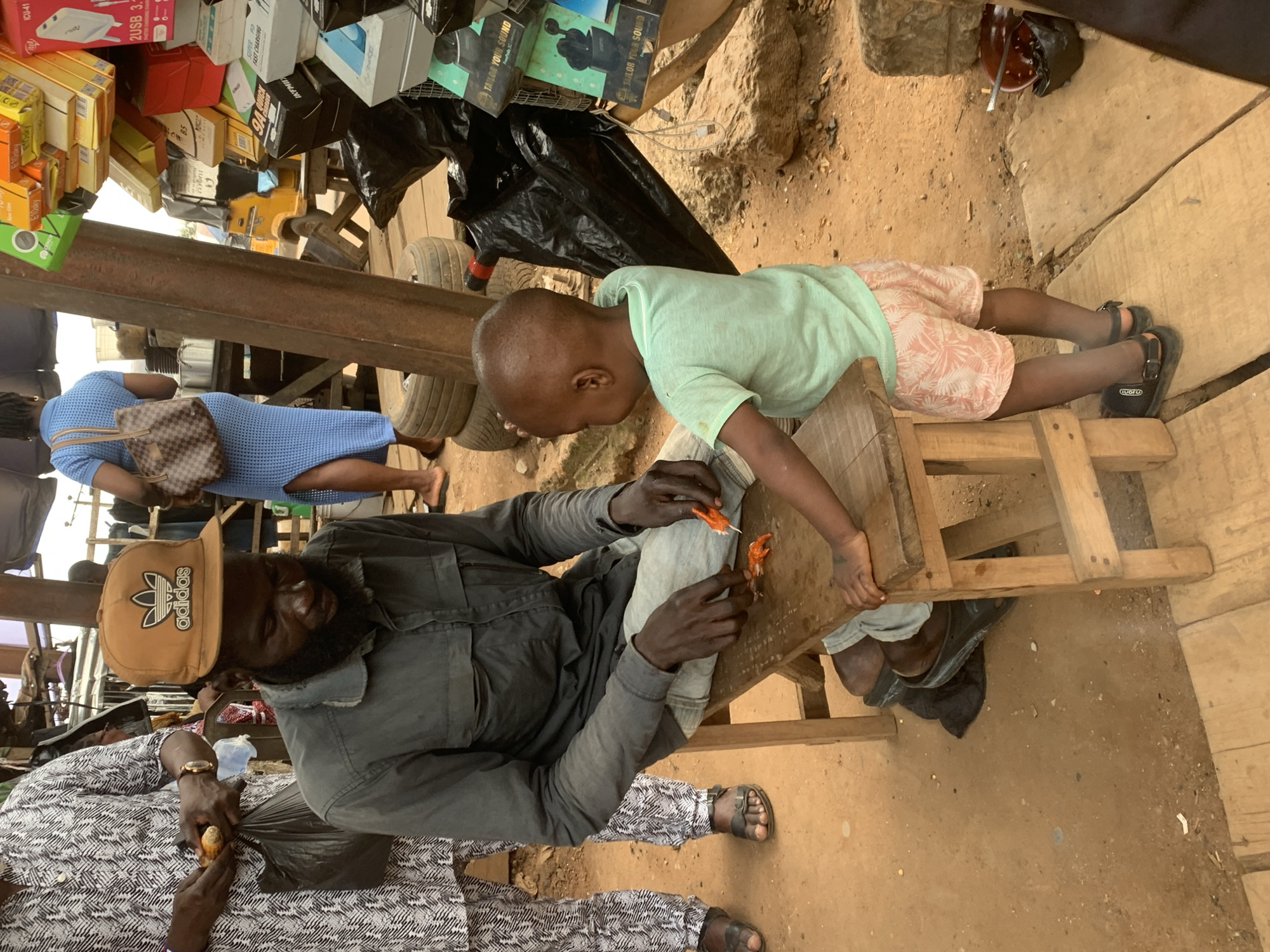 The image depicts a scene at an outdoor market in Shagamu, Nigeria. A Black man with a beard and wearing a grey shirt and light-colored pants sits on a wooden stool, looking down at something in his hands. He is holding a small orange object and a small blade, possibly preparing food or a craft. Beside him, a young Black child, facing away from the camera, stands and looks towards the man. The child wears a light green t-shirt and pink patterned shorts.
The environment appears to be a bustling marketplace. In the background, there are various stalls with goods displayed, including boxed electronics and what appears to be food items. Some tires are visible near the stalls. The ground is dirt, with some wooden planks laid out in the foreground. The lighting suggests it is daytime, likely overcast or in the shade of market structures. The overall impression is one of everyday commerce and activity in a Nigerian market.