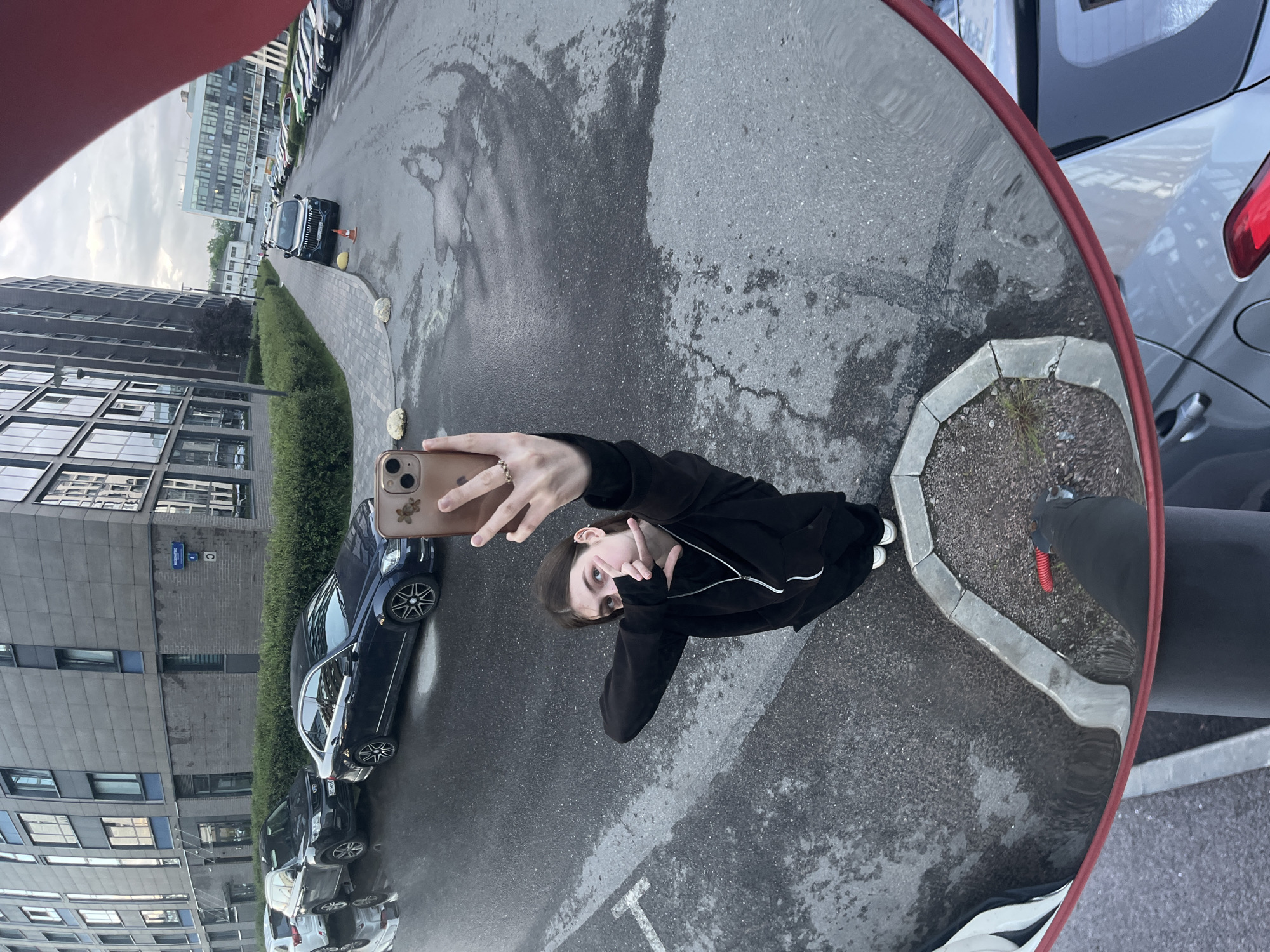 A young woman is taking a selfie in a convex mirror, standing on a wet asphalt street in front of a modern apartment building. She is wearing a black hoodie and white sneakers. Her left hand is holding a smartphone, and her right hand is positioned near her face, forming a V-shape with her fingers. In the background, several cars are parked along the street and sidewalk. The scene appears to be in a residential area of a city.