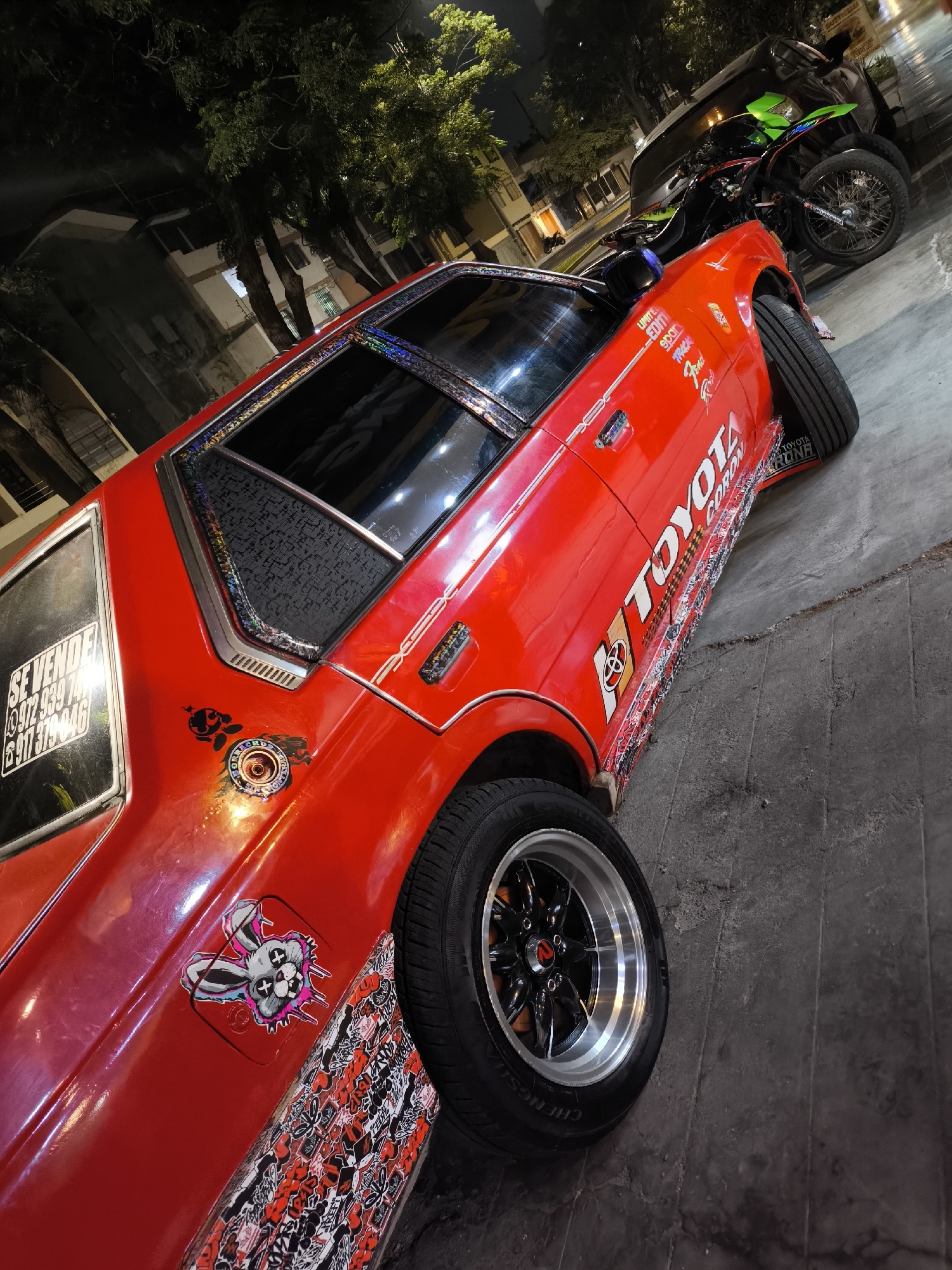 The image shows a red Toyota Corolla parked on a city street at night. The car has custom decals and stickers, including a "Se Vende" (For Sale) sign in the rear window with a phone number. A motorcycle is parked behind the car. The street appears to be in a residential area.
