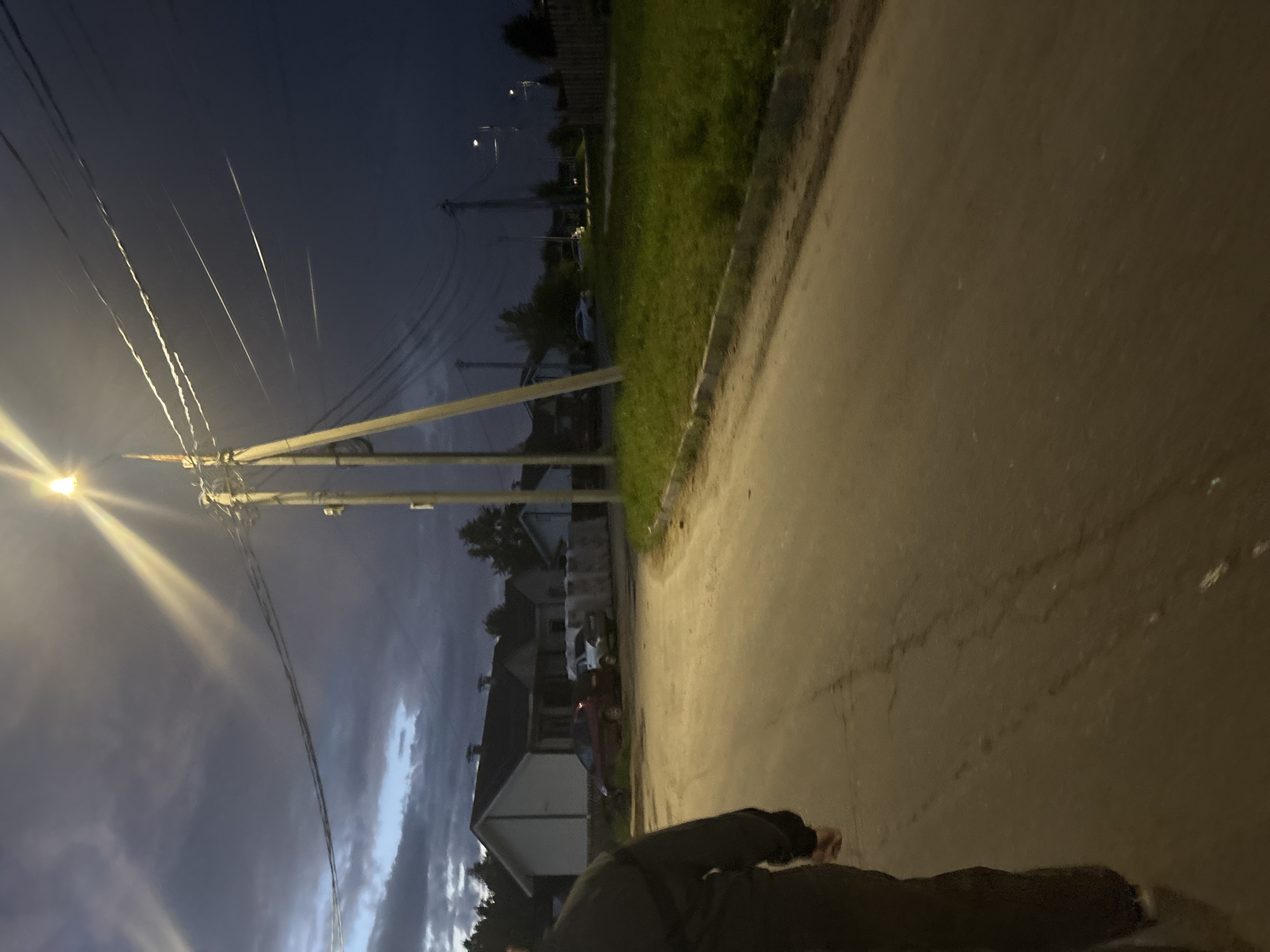 The image, captured from a tilted perspective, depicts an outdoor residential street scene at nighttime or late dusk. In the lower right foreground, a partial view of a person is visible, dressed in dark clothing, standing or walking on a paved road or path. This paved surface, which shows some cracks and wear, runs along the right side of the frame and is bordered by a narrow strip of green grass.
A prominent utility pole stands on the left side of the grass strip, featuring a brightly lit street lamp that emits a strong glare and beams of light. These beams illuminate a complex network of power lines extending from the pole and across the dark, cloudy sky. The sky itself is a deep blue-grey with visible cloud formations, indicating an overcast night.
In the background, behind the utility pole and power lines, several dimly lit residential houses with pitched roofs and at least two parked cars are discernible. The overall lighting is dim, with the street lamp serving as the primary light source, creating distinct contrasts between the illuminated areas and the surrounding darkness. There is no visible text in the image.