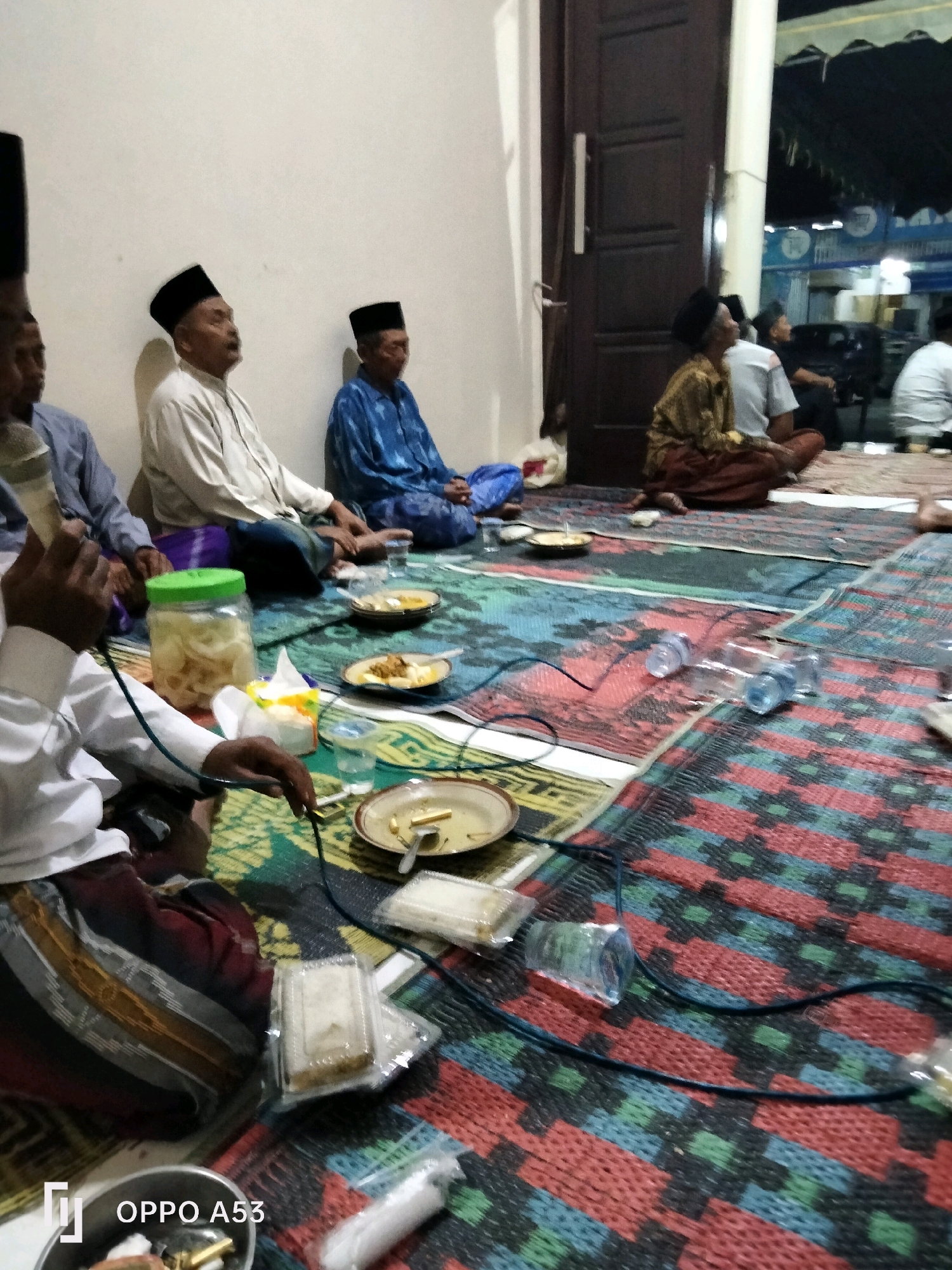 The image depicts a gathering of men, likely Indonesian, seated on woven mats on the floor, engaged in a meal. Most are wearing traditional Islamic attire, including peci hats and sarongs. One man in the foreground is holding a microphone. Plates with food, glasses of water, and a jar of pickled vegetables are visible on the mats. In the background, a doorway leads to an outdoor area with a tent or canopy, suggesting an event is taking place. The overall setting appears to be a modest, possibly communal space, with a light-colored wall and a dark wooden door. The presence of the "OPPO A53" watermark in the lower-left corner indicates the image was taken with that specific phone model. The context of the gathering, the attire, and the general atmosphere suggest a religious or cultural event, potentially in a village or community setting. Without further information, it's impossible to pinpoint the exact location within Trenggalek, Indonesia.