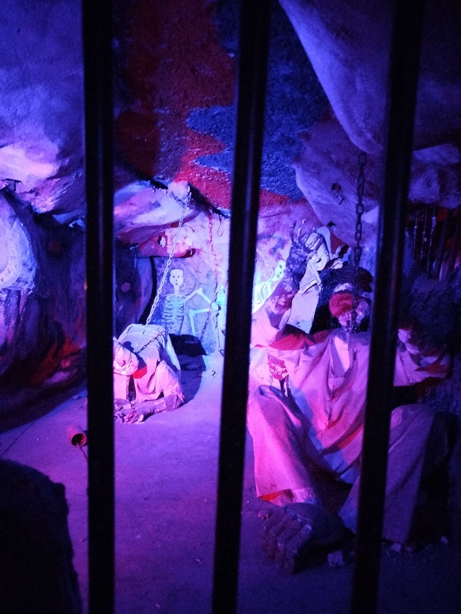 The image, seemingly taken in Xuân Thành, Vietnam, depicts a staged dungeon-like setting viewed through black metal bars, likely part of a tourist attraction or themed venue. The lighting is predominantly blue and pink, creating a spooky atmosphere. Inside the enclosure, there are several figures or mannequins dressed in tattered clothing. One figure appears to be kneeling, with another seemingly chained and slumped over. A skeleton hangs in the background. The environment appears to be an artificial cave with a textured and uneven surface painted with red and blue patches.