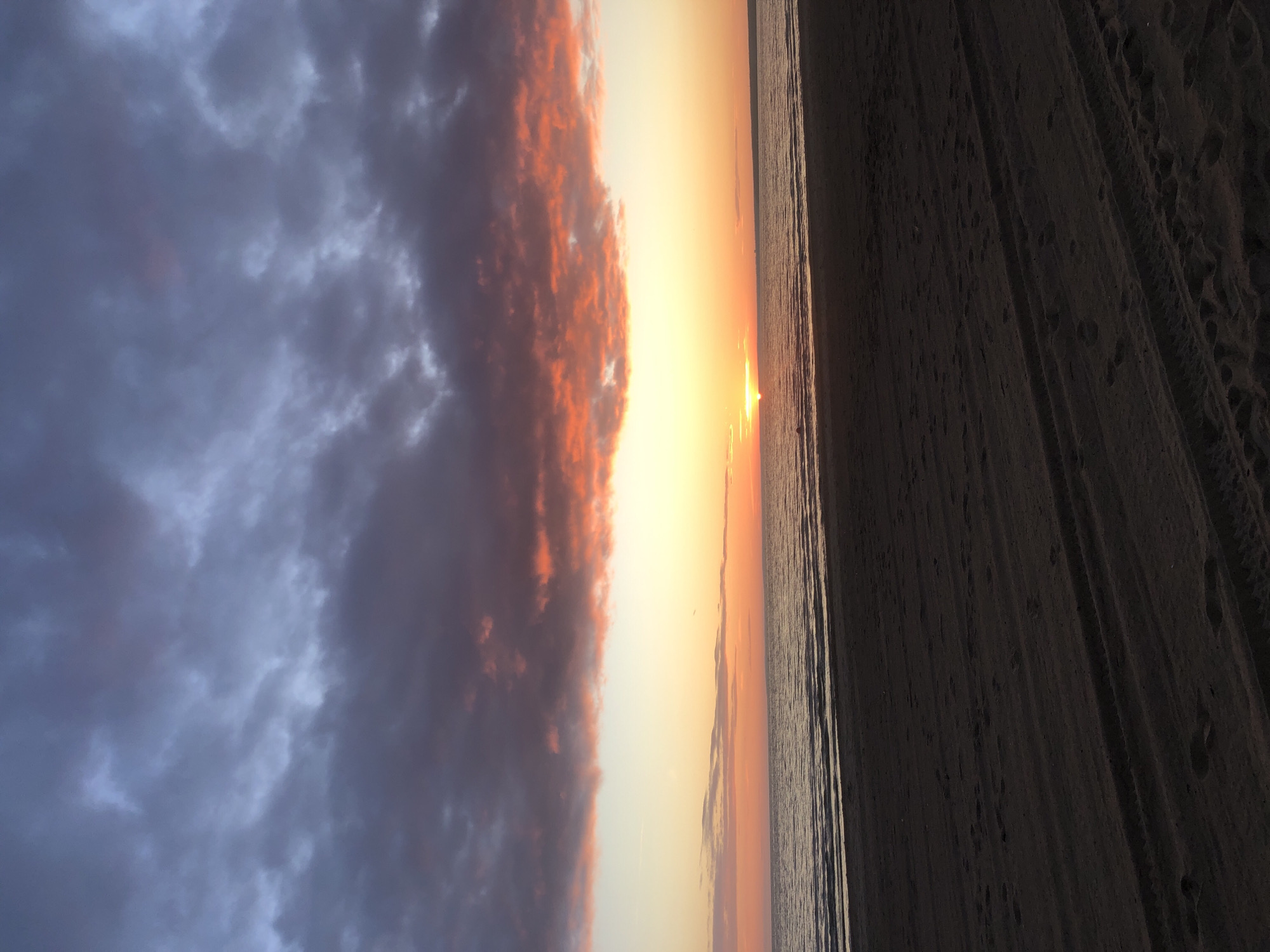 The image shows a sunset over the Gulf of Finland, viewed from the beach in Sestroretsk, Russia. The sky is filled with a dramatic display of clouds, ranging in color from deep grays and purples at the top to vibrant oranges and yellows near the horizon. The sun is a bright, small orb just above the water, casting a warm glow. The water is calm, with gentle waves lapping at the shore. The foreground is the dark sand of the beach, showing tire tracks and footprints. The overall scene is serene and picturesque, characteristic of a sunset in this coastal town.
