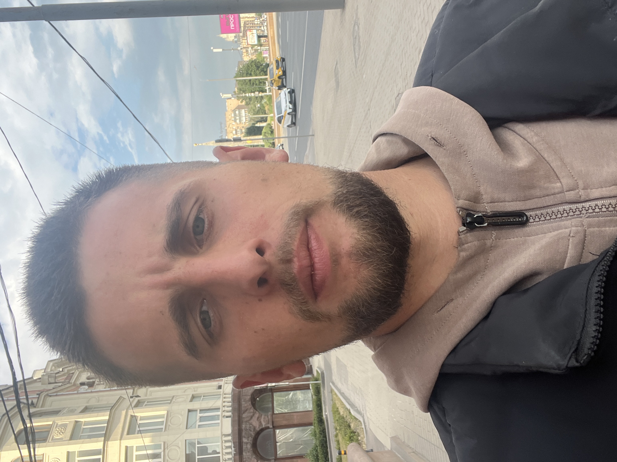 A man with short brown hair and a beard is standing on a sidewalk in front of a building with a classic facade. He is wearing a black jacket over a beige hoodie. In the background, there is a street with cars and a billboard with Russian text that reads "ВЫТЯЖКА ПРО" which likely refers to a ventilation system. The sky is partly cloudy, and the sun is casting shadows. The overall scene depicts a casual outdoor setting in an urban environment.