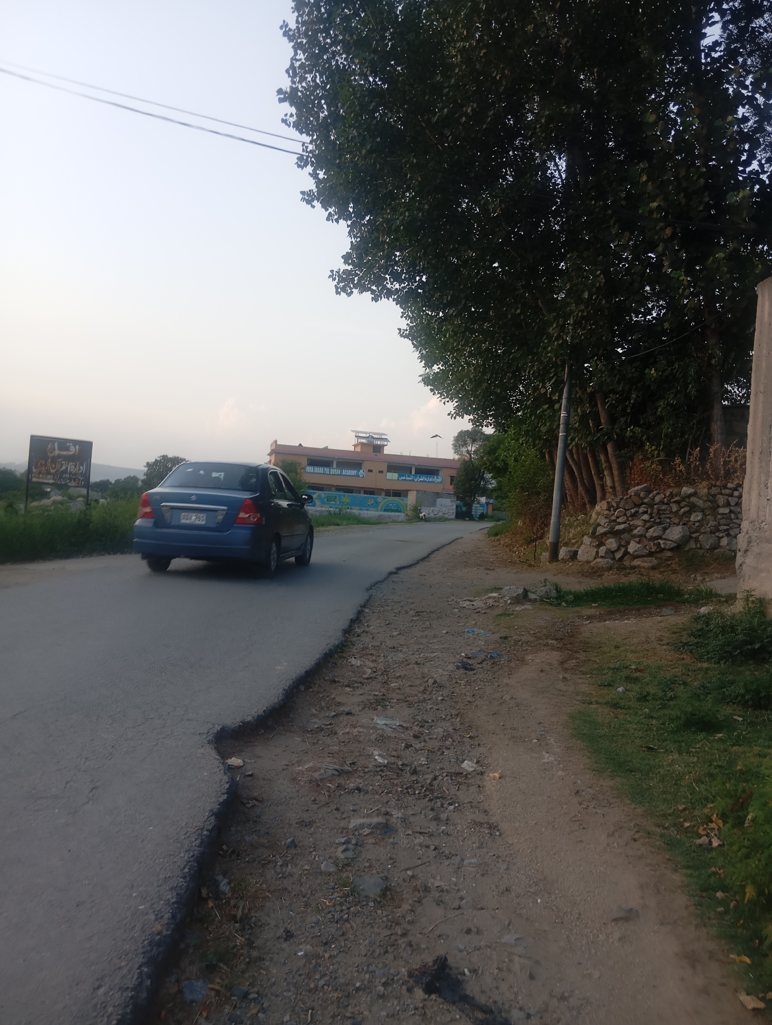 A blue sedan travels down a paved road in an outdoor setting. The car is in motion, seen from the rear left quarter. The road is cracked and appears to be in disrepair on the right side, transitioning to a dirt path. To the left of the road, a sign with text in a foreign language is visible, along with tall green grass and shrubs. In the background, a multi-story building with a beige facade and a dark roof stands, with signs of an educational institution displayed. Trees with dense green foliage are prominent on the right side of the image, and a stone wall is also present. Power lines are visible in the sky. The lighting suggests it is daytime, possibly late afternoon or early morning, with a hazy sky.