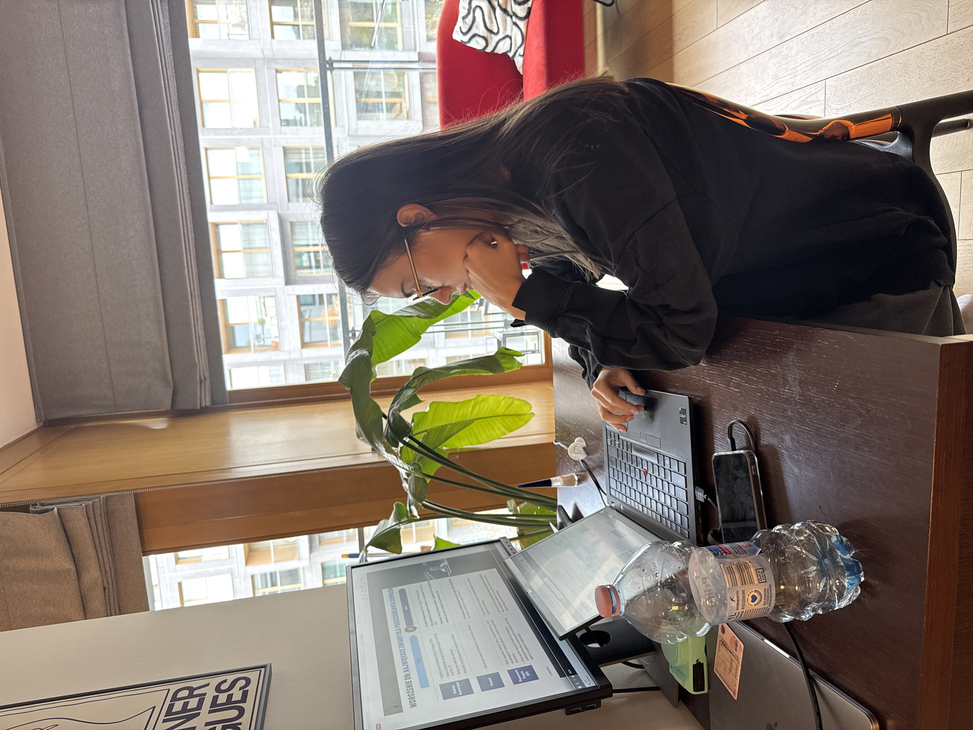 An eye-level, wide shot captures a person working intently at a desk, illuminated by natural light from a large window. The scene is set in what appears to be a modern home office or workspace in Warsaw, Poland.
The primary subject is a young woman with long, dark hair, wearing glasses, a black long-sleeved top, and grey sweatpants. She is seated at a dark wooden desk, her body turned slightly to the left, leaning forward with her right hand resting near her face, suggesting deep focus or contemplation. Her left hand is on the trackpad of a black laptop.
On the desk, besides the laptop, an external monitor is prominently displayed, showing a document or presentation. A transparent plastic water bottle, partially full, sits next to it. A black smartphone, a pen, and white earbuds are also visible on the desk. A large, leafy green plant, possibly a bird of paradise, is situated between the woman and the monitors. In the background, a vibrant red armchair with a white patterned cushion adds a splash of color.
Beyond the desk, a large window spans the background, offering a view of a modern building with numerous square-paned windows, indicating an urban setting. Beige curtains or blinds frame the window. The room features light-colored wooden flooring and walls.
Visible text on the external monitor reads: "WDROŻENIE 09: NAJWIĘKSZE ZMIANY DLA POSZCZEGÓLNYCH ZESPOŁÓ" (IMPLEMENTATION 09: THE BIGGEST CHANGES FOR INDIVIDUAL TEAMS), along with other words like "Funkcje," "Baycry and," "K," and "MEGAZOWANA," likely part of the content being displayed. A partially visible poster or artwork below the desk shows the text "VER SUES." Text on the water bottle label is also visible, including "BULING" and other product details. The time of day appears to be daytime, with bright ambient light filling the room.