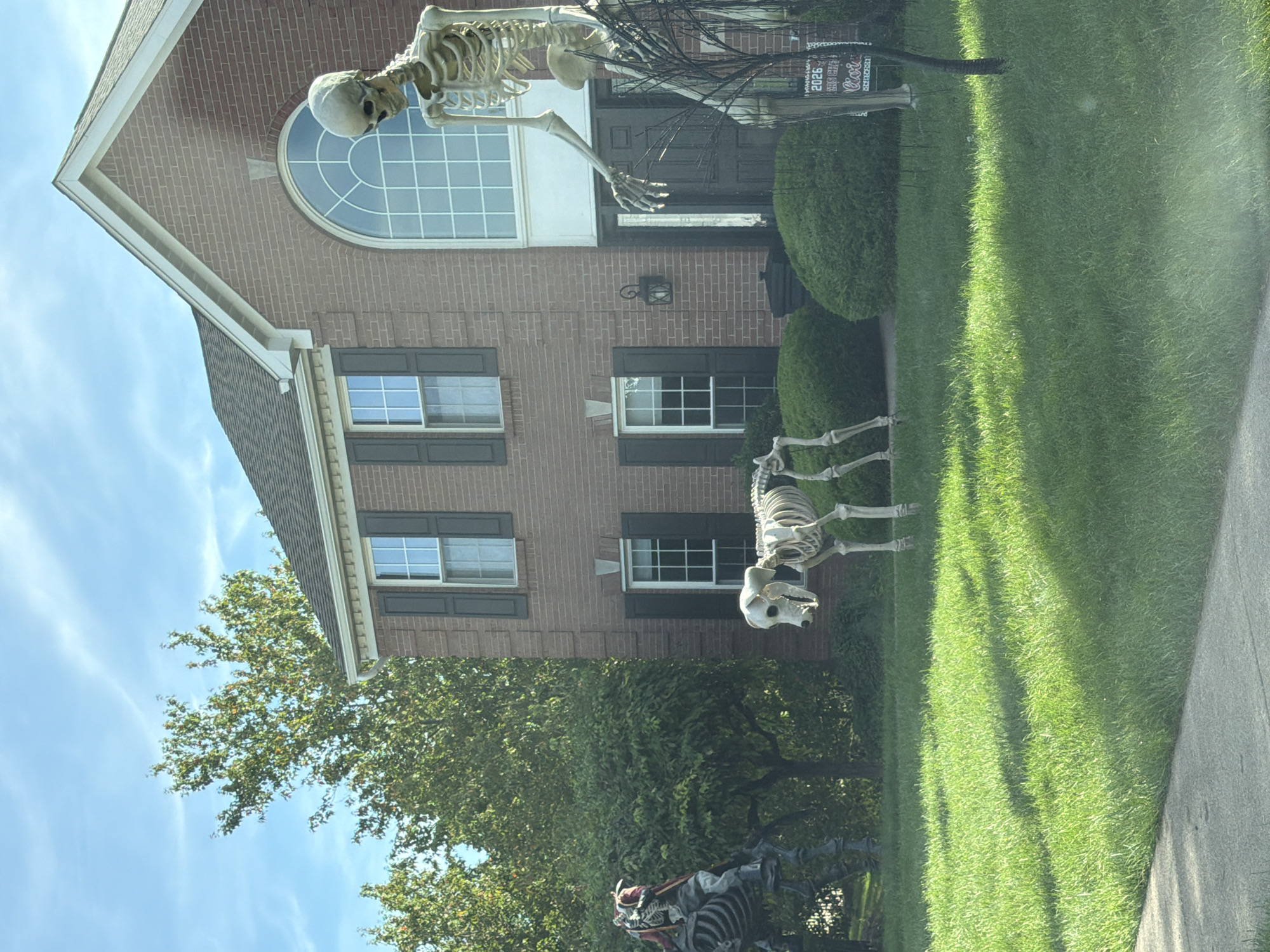In Peters Township, United States of America, a two-story brick house with a dark roof and arched window above the front door is decorated for Halloween. A large skeleton stands on the right side of the lawn, reaching its arm out. To the left of the house, a skeleton dog is positioned on the grass. Further to the left, a skeleton rider on a skeletal horse is visible. The lawn is manicured, and manicured bushes are placed in front of the house. The sky is clear and blue with some wispy clouds.