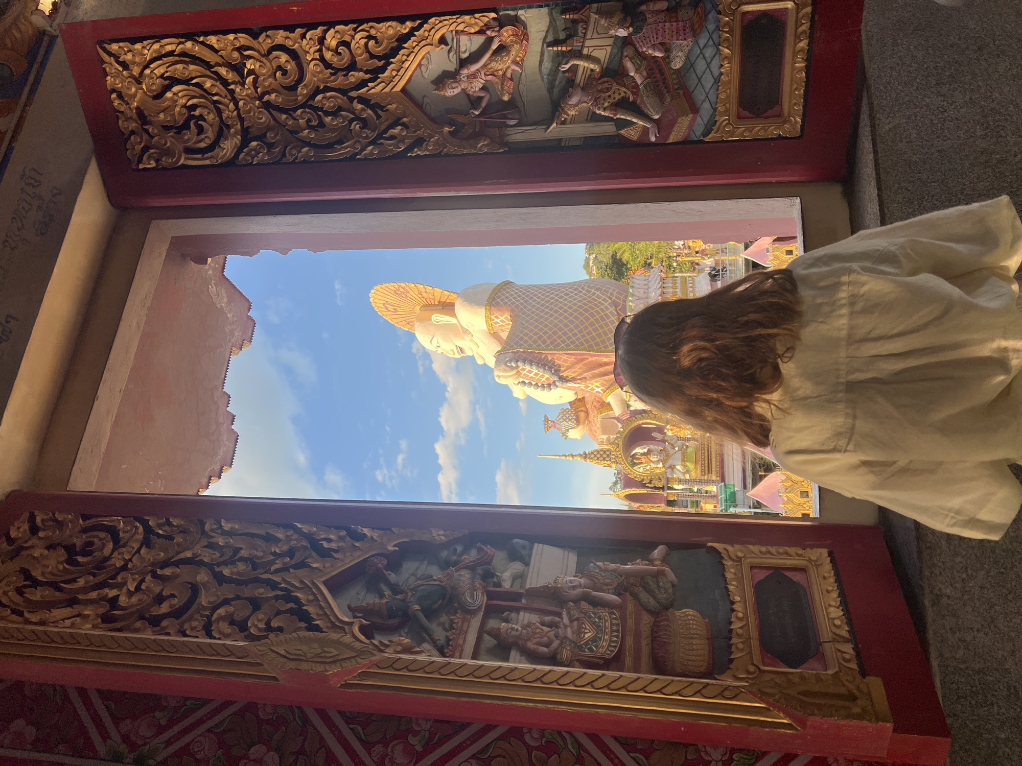 The image captures a view through an ornate, red-framed opening, likely a doorway or window of a temple. The opening is decorated with intricate golden carvings of figures and patterns. Through this opening, a person, seen from behind, stands and looks out. They are wearing a light-colored, possibly cream or off-white, garment with a gathered neckline. Their long, brown hair is visible.
The scene beyond the opening is dominated by a massive, reclining statue of Buddha, appearing to be made of a pale material with golden accents. The statue is adorned with a decorative garment and a wide-brimmed, golden hat or halo. The Buddha statue is set against a bright blue sky with scattered white clouds, suggesting a clear, daytime setting. In the background, more of the temple complex is visible, with golden spires and structures. The overall atmosphere appears peaceful and awe-inspiring, with the grand scale of the Buddha statue conveying a sense of reverence. The time of day is likely daytime, given the bright sky. The location is suggested to be Baan Plai Laem, Thailand, which aligns with the architectural style and religious imagery. No visible text is present in the image.