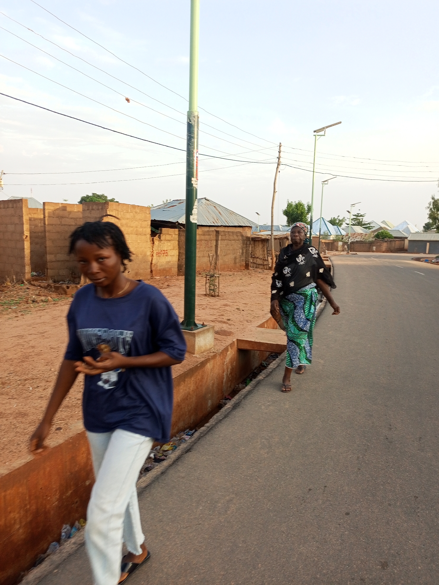 Two women walk along a paved road beside a dirt area with brick structures, likely in a settlement or neighborhood. The woman in the foreground is younger, wearing a blue t-shirt and white pants. The other woman is older, wearing a black top and a green and blue patterned skirt. They are both wearing sandals. Street lights are visible.